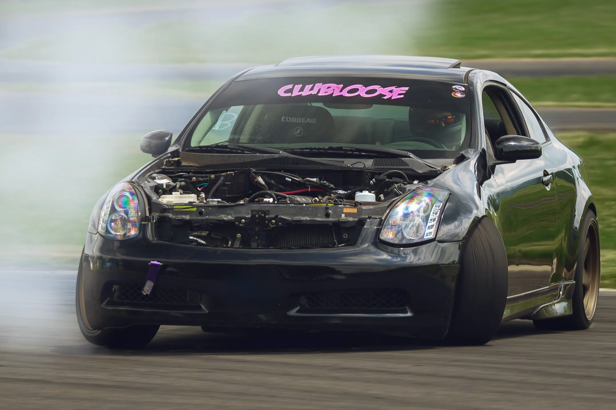 Black drift car with exposed engine bay and no front bumper drifting on a racetrack, creating tire smoke, with a driver wearing a helmet inside.