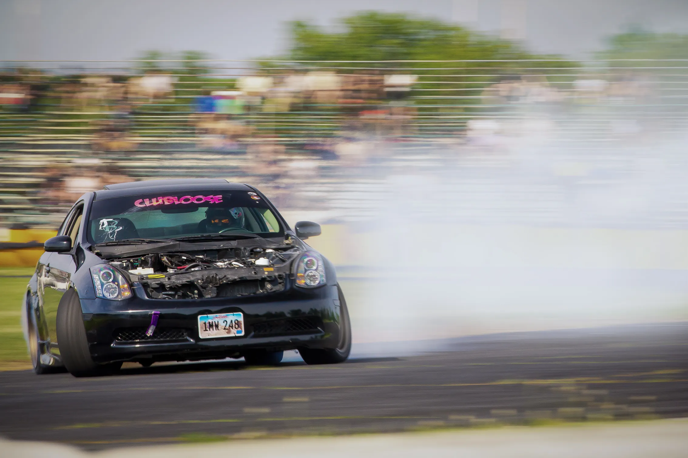 Black car with exposed engine drifting on a race track, producing smoke from rear tires, with blurred spectators in the background.
