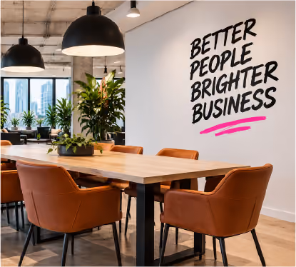 A modern office meeting area with a wooden table, brown chairs, hanging black pendant lights, and a wall graphic reading “BETTER PEOPLE BRIGHTER BUSINESS.”