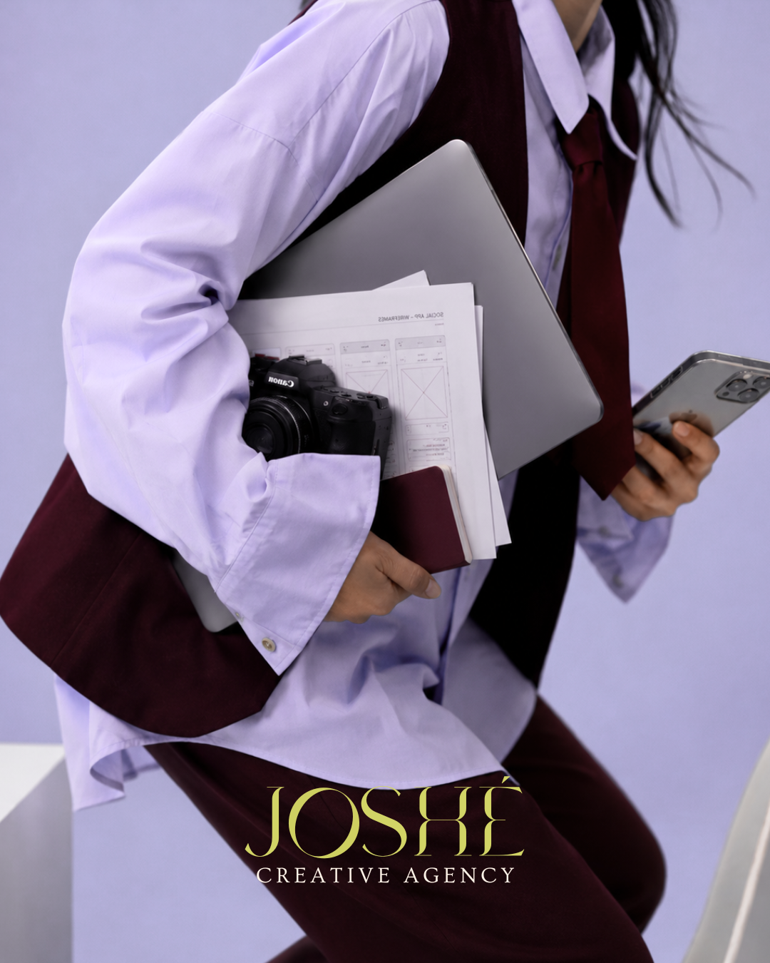 Henkilö, jolla on violetti kauluspaita, viininpunainen liivi ja solmio, pitelee kannettavaa tietokonetta, paperidokumentteja ja kameraa käsivarrellaan ja selaa älypuhelinta.