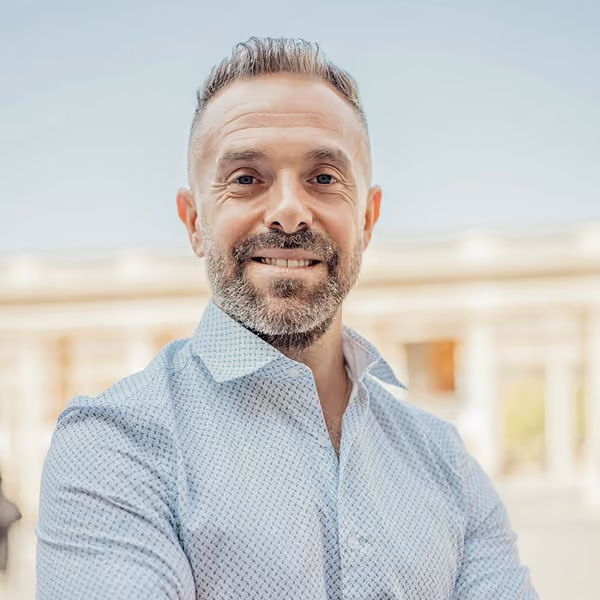 Smiling middle-aged man with short hair and beard wearing a patterned light shirt outdoors.