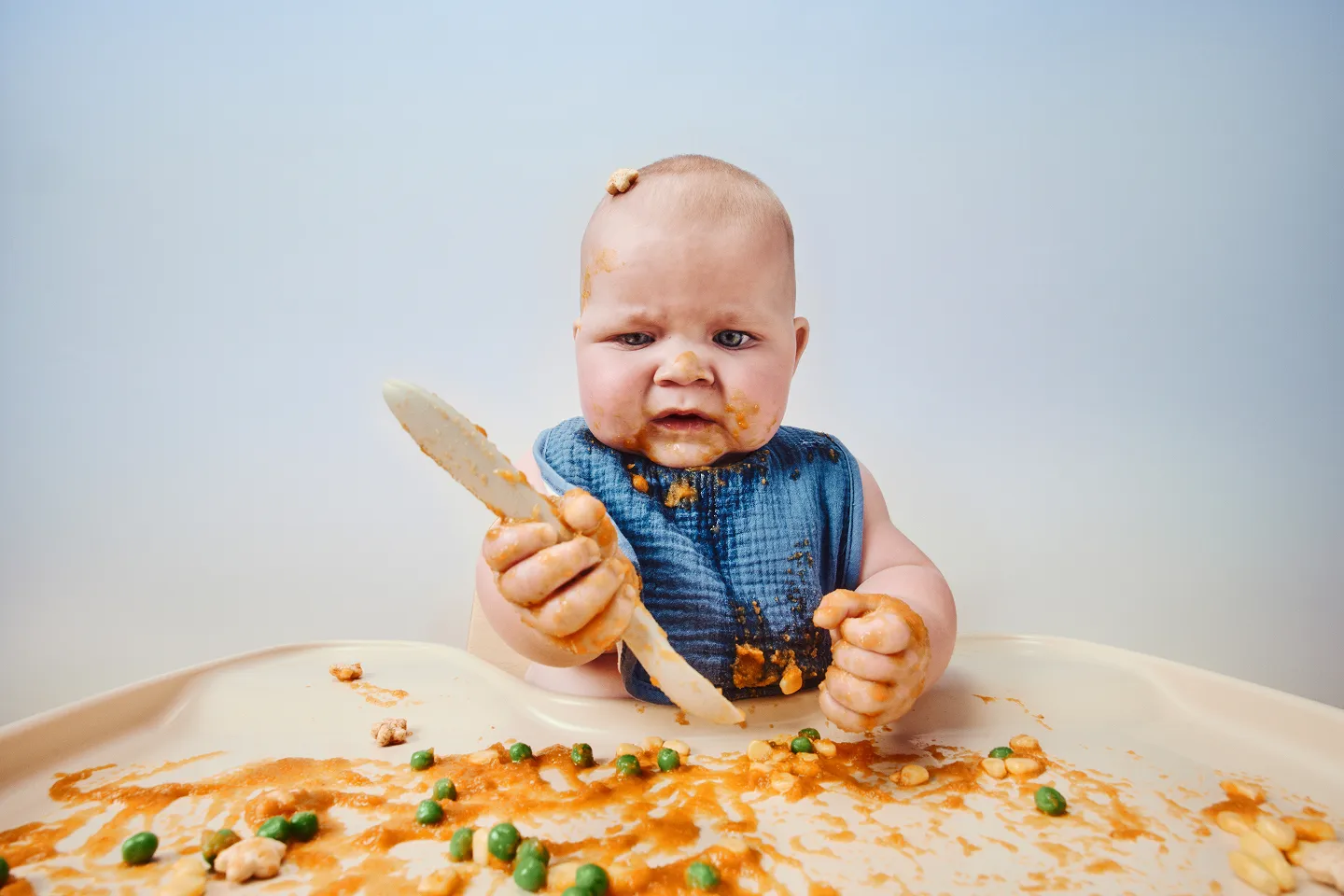 Baby in a blue bib at a high chair holding a wooden spoon, with a messy tray of food in front of them