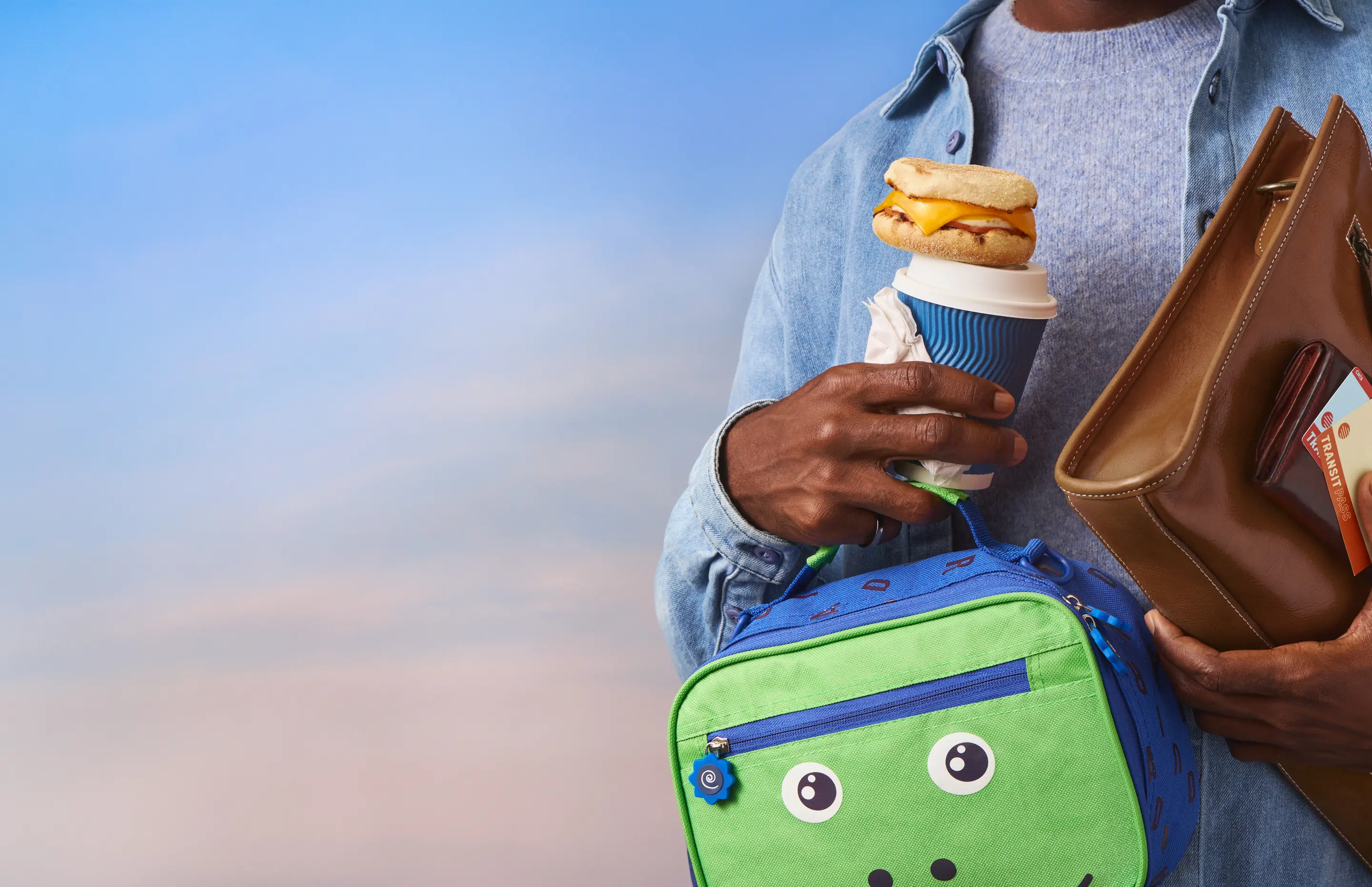 Person holding a coffee cup topped with a breakfast sandwich, a kids' lunch bag and a brown tote