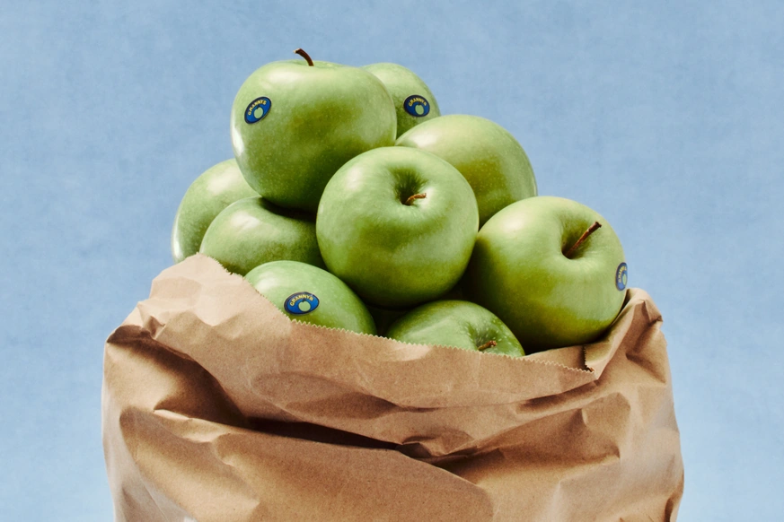 Paper grocery bag full of green apples against a pale blue background
