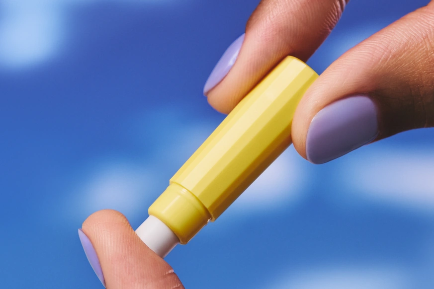 Two hands with lilac nails holding a yellow injection pen against a blue sky