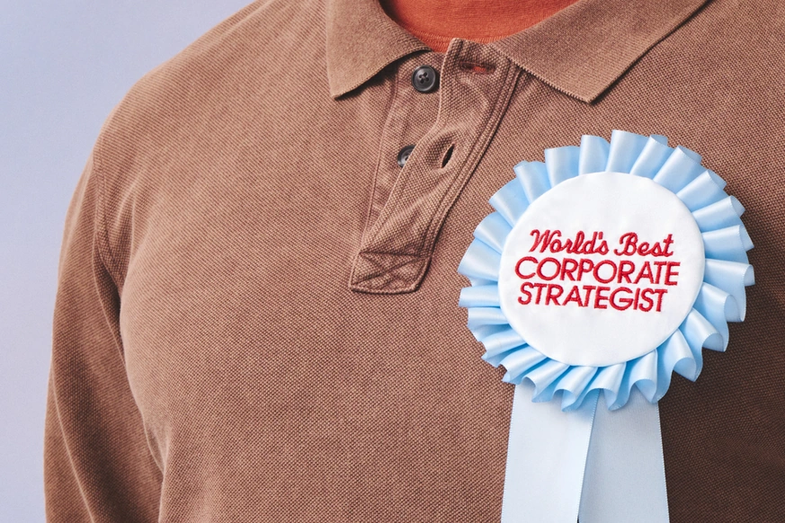 Person in a brown polo shirt wearing a World's Best Corporate Strategist rosette