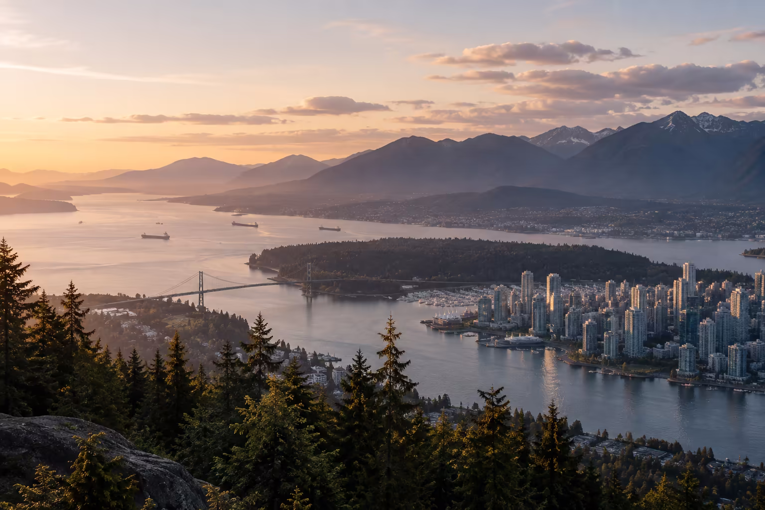 Sunset view over a coastal city with dense high-rise buildings, surrounded by water, forested areas, and mountains in the background, with several ships in the water and a prominent bridge connecting land sections.