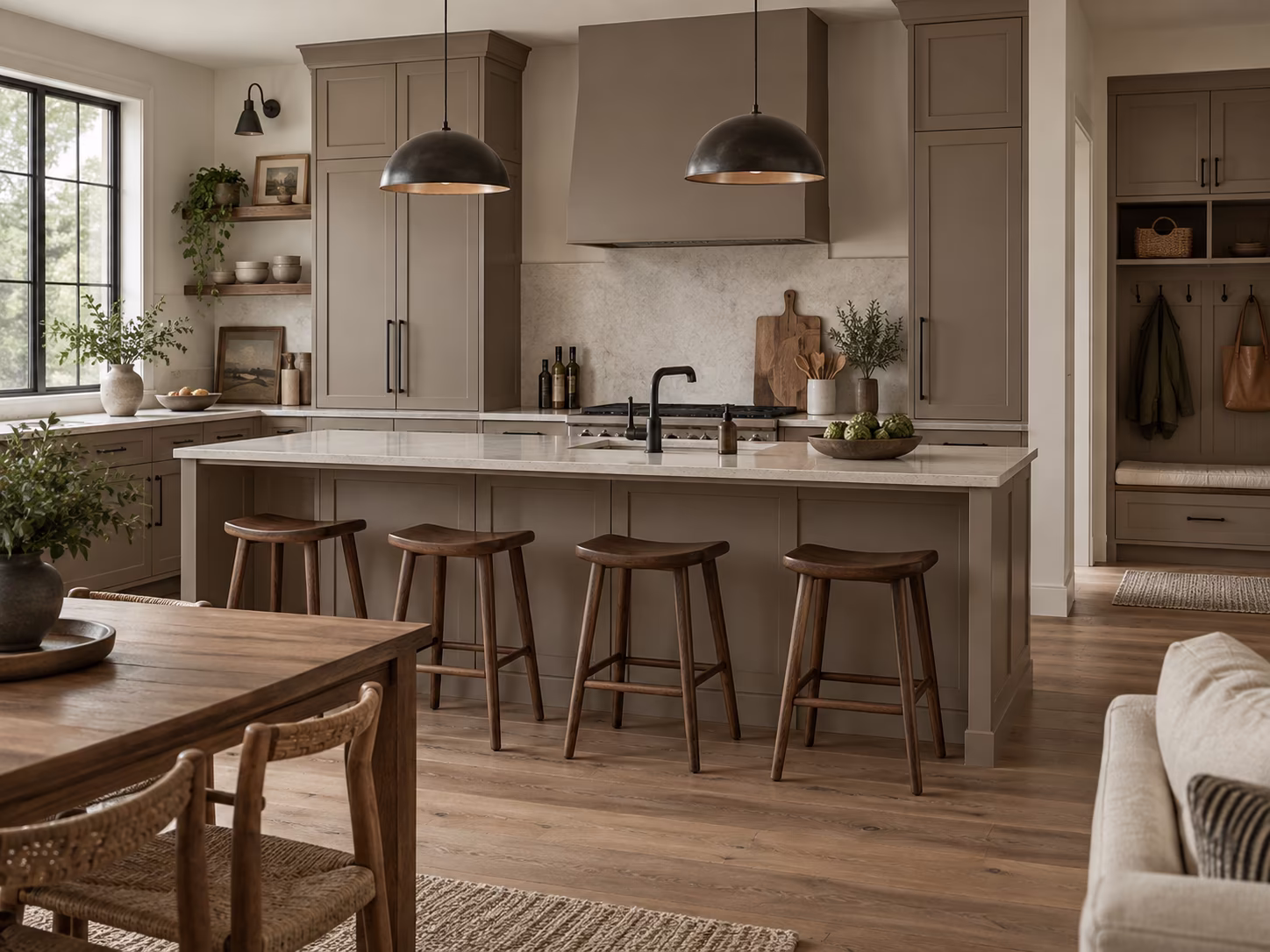 Modern kitchen with light brown cabinetry and a large island featuring a white countertop and four wooden stools, two dark pendant lights hang above, wooden dining table and chairs in the foreground, plants and decorative items on counters and shelves.