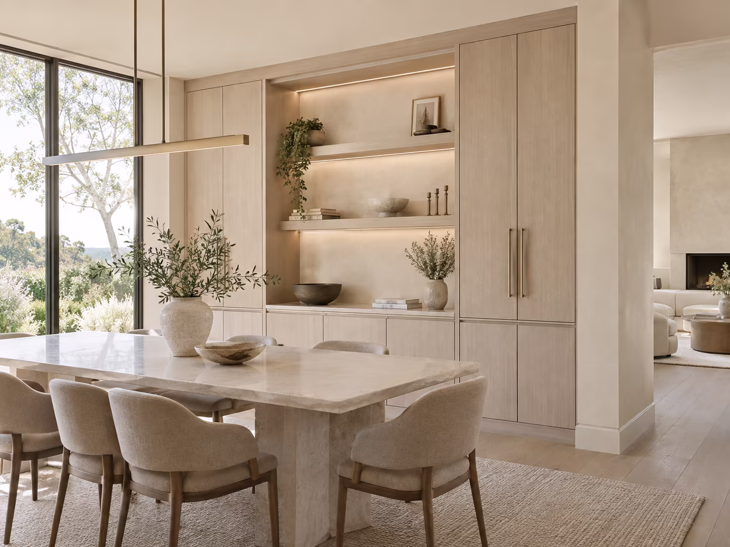 Modern dining room with a large rectangular marble table surrounded by six beige upholstered chairs, a built-in light wood cabinet with open shelves displaying decorative items, a large window showing trees outside, and a beige carpet underneath the table.