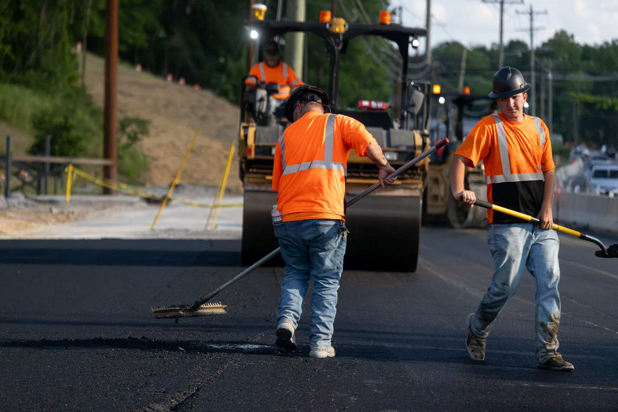 Construction workers in safety vests and helmets conversing at an asphalt paving site with equipment in the background.