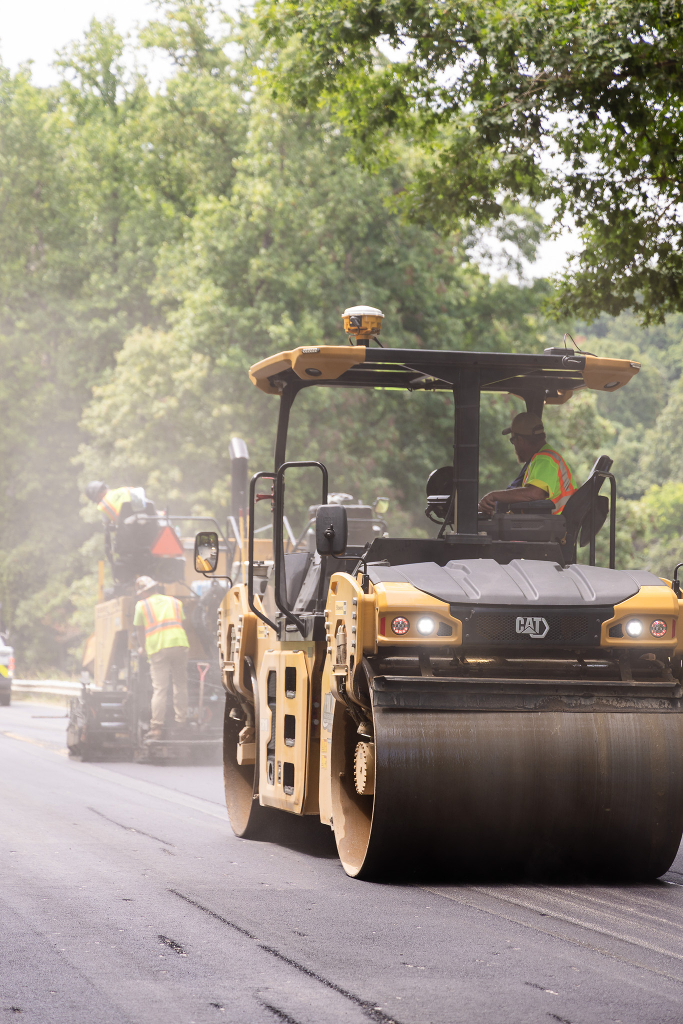 Construction workers in safety vests and helmets spreading fresh asphalt with machinery and tools on a parking lot near a fenced sports court.