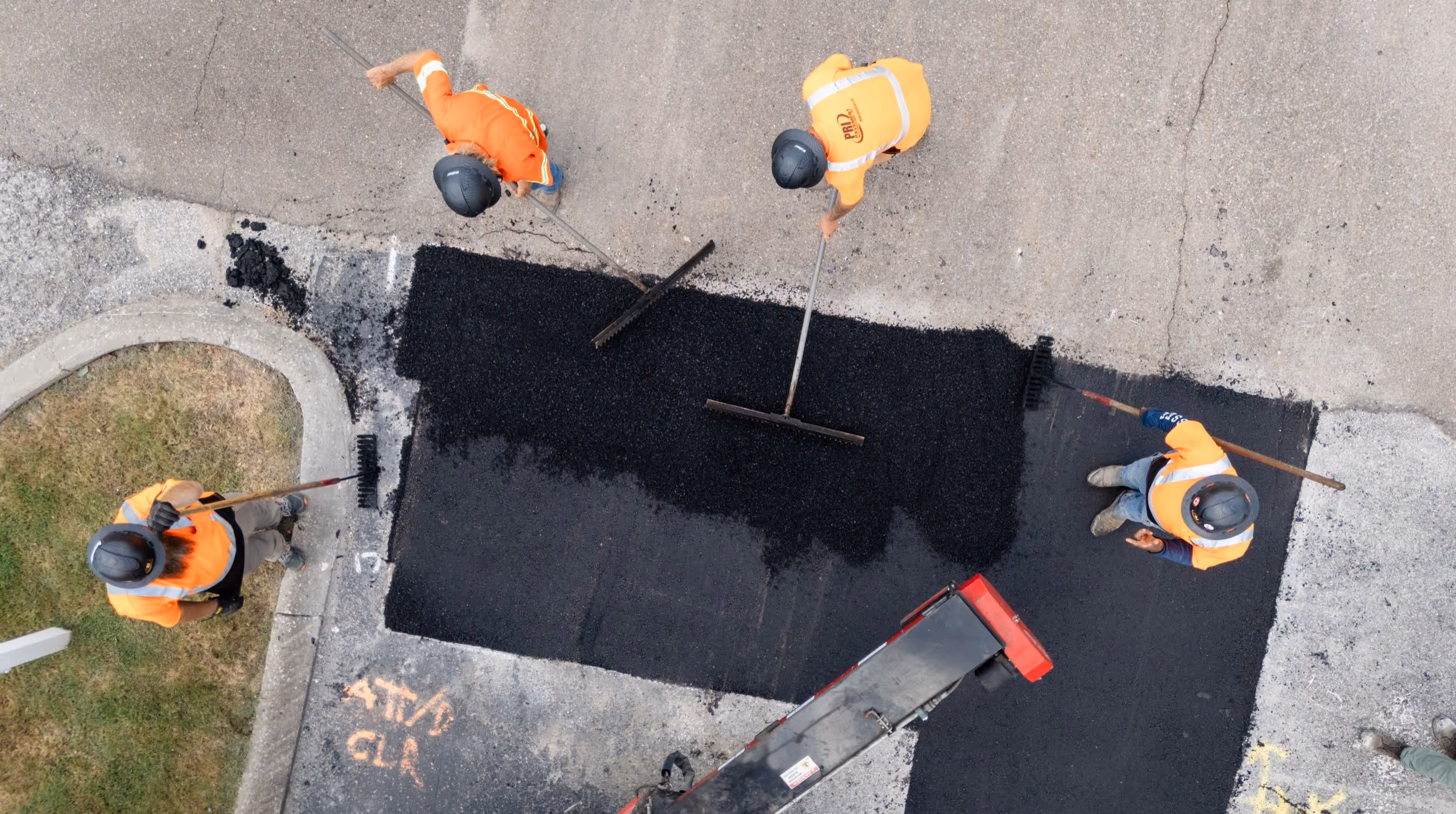Aerial view of four construction workers in orange safety vests and helmets spreading fresh asphalt on a road patch.