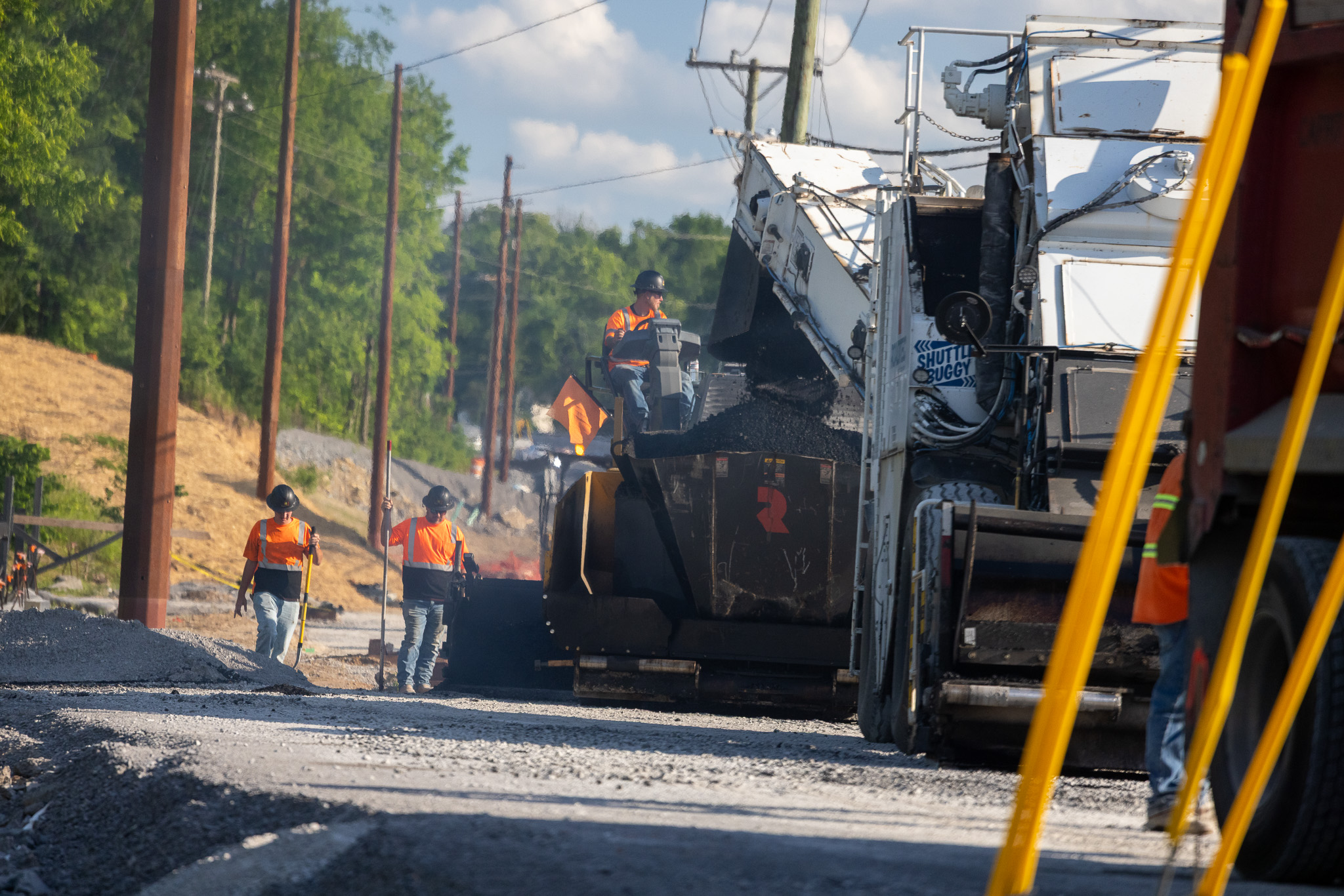 Construction workers wearing safety vests and hard hats paving a road with asphalt using a broom and paving machine.