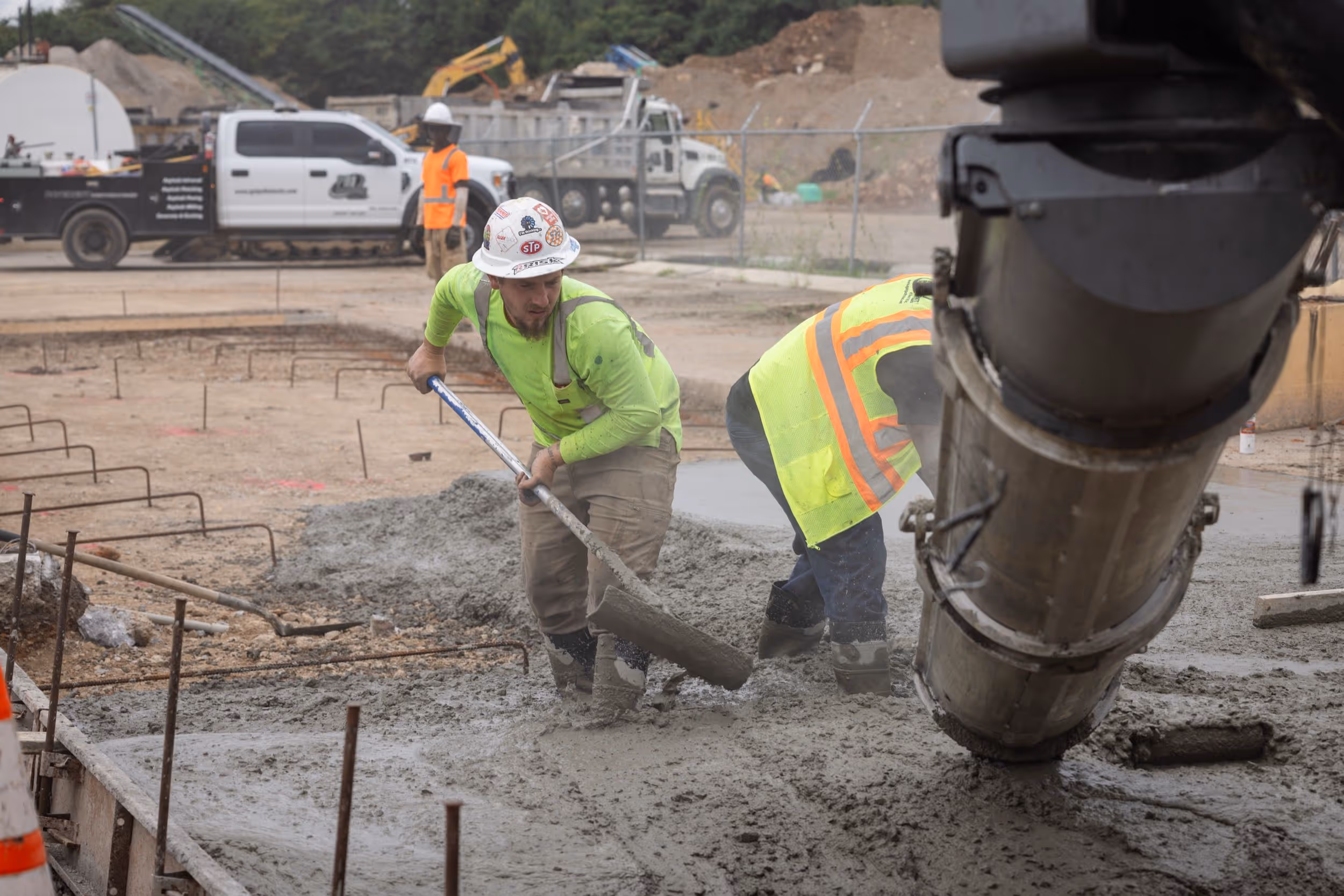 Two construction workers in fluorescent safety gear pouring and spreading wet concrete at a construction site.
