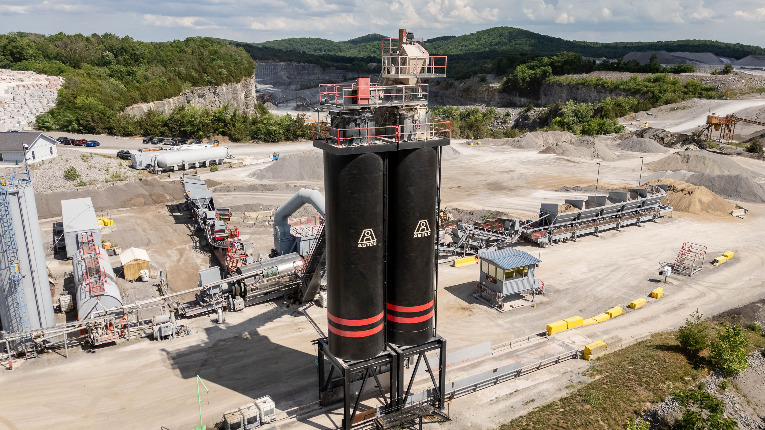Industrial asphalt plant with tall black storage silo labeled PRI Pavement Restorations and Astec, conveyor belt, and various construction vehicles outdoors.