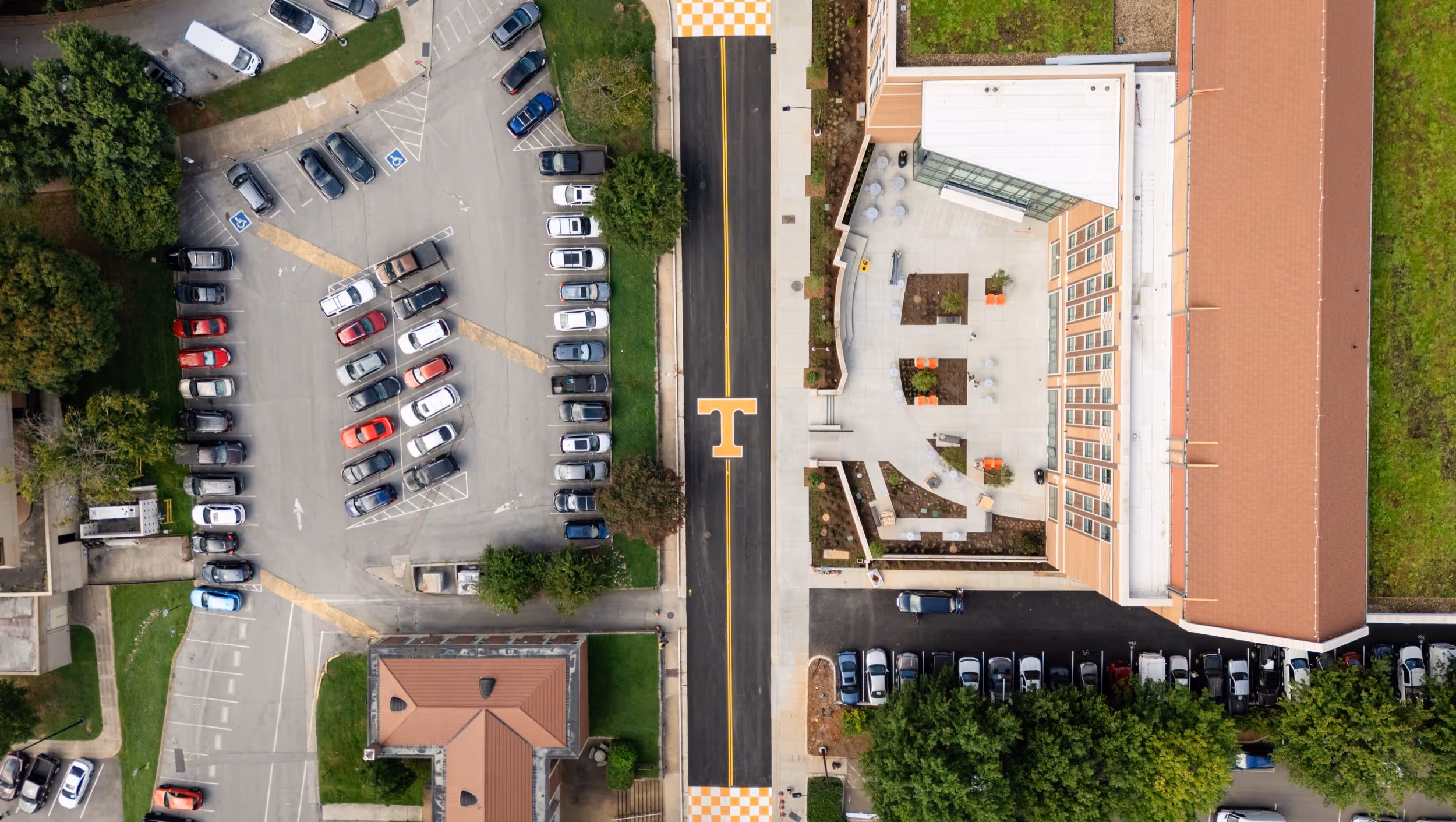 Aerial view of a parking lot with numerous parked cars next to a modern building with outdoor seating and a black road with an orange 'T' symbol in the center.
