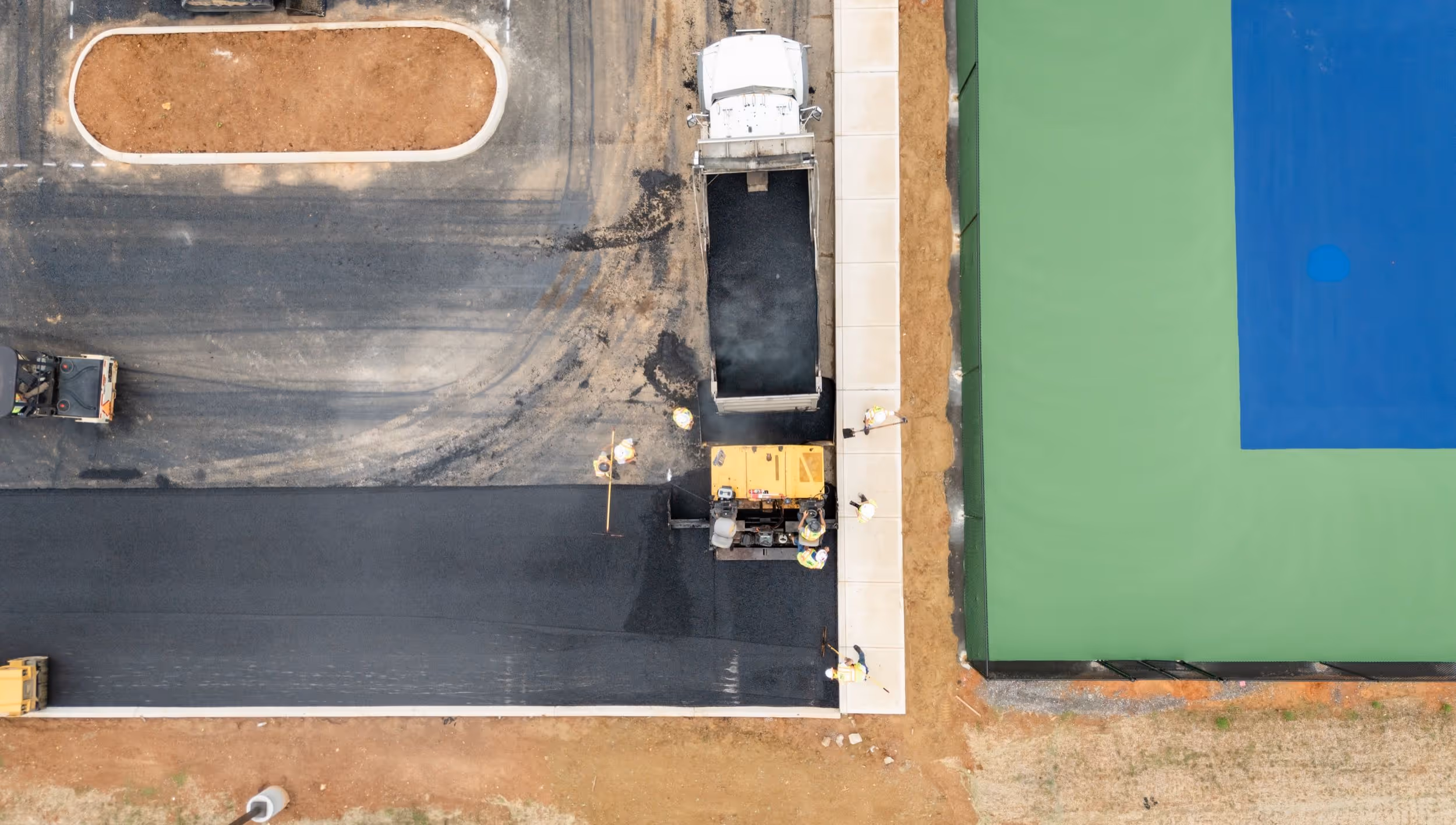 Aerial view of workers paving a black asphalt road next to a green and blue tennis court.