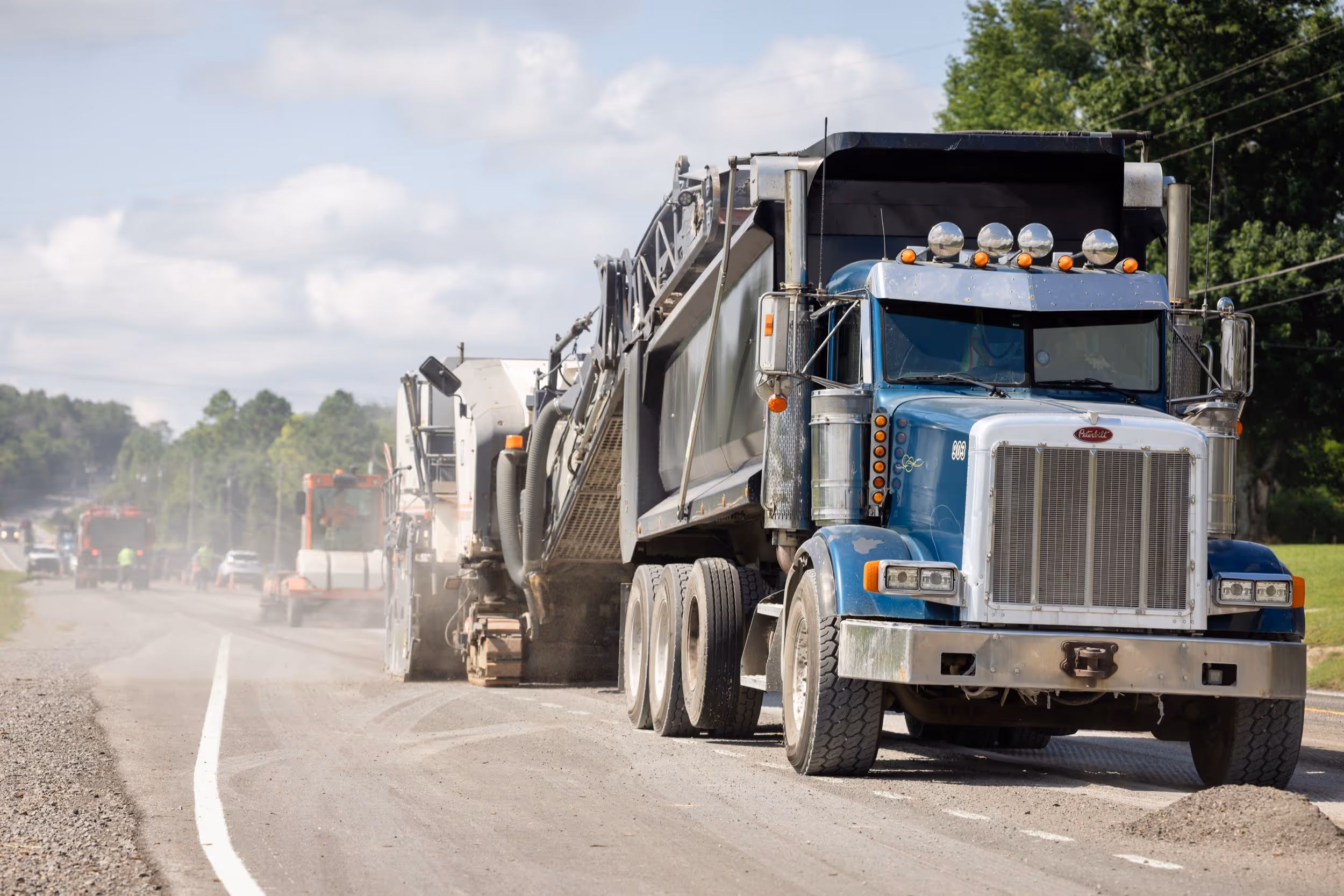 Blue dump truck positioned behind road construction machinery on a gravel road with workers and vehicles in the background.