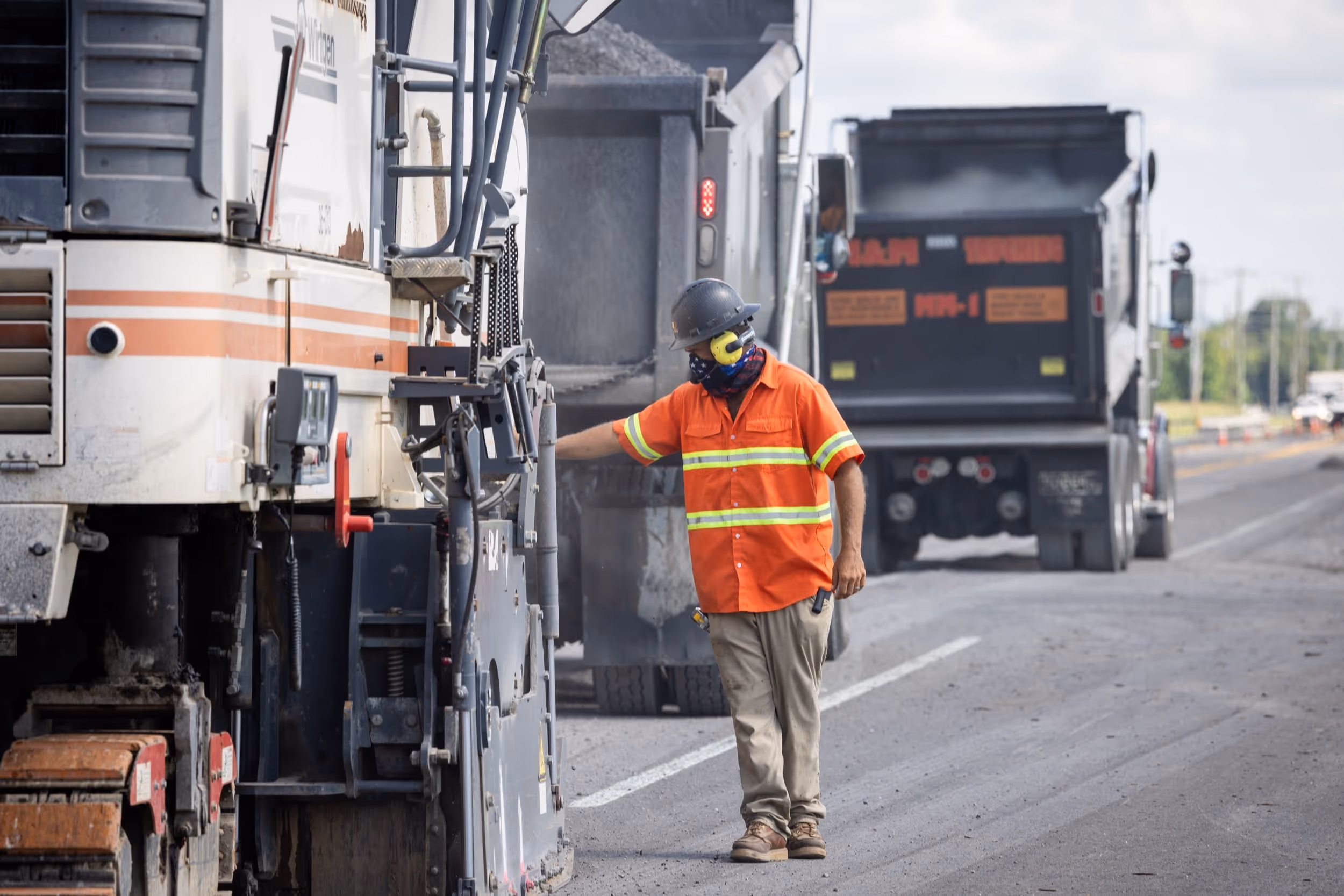 Construction worker in orange high-visibility clothing and protective gear operating road paving machinery on a highway.