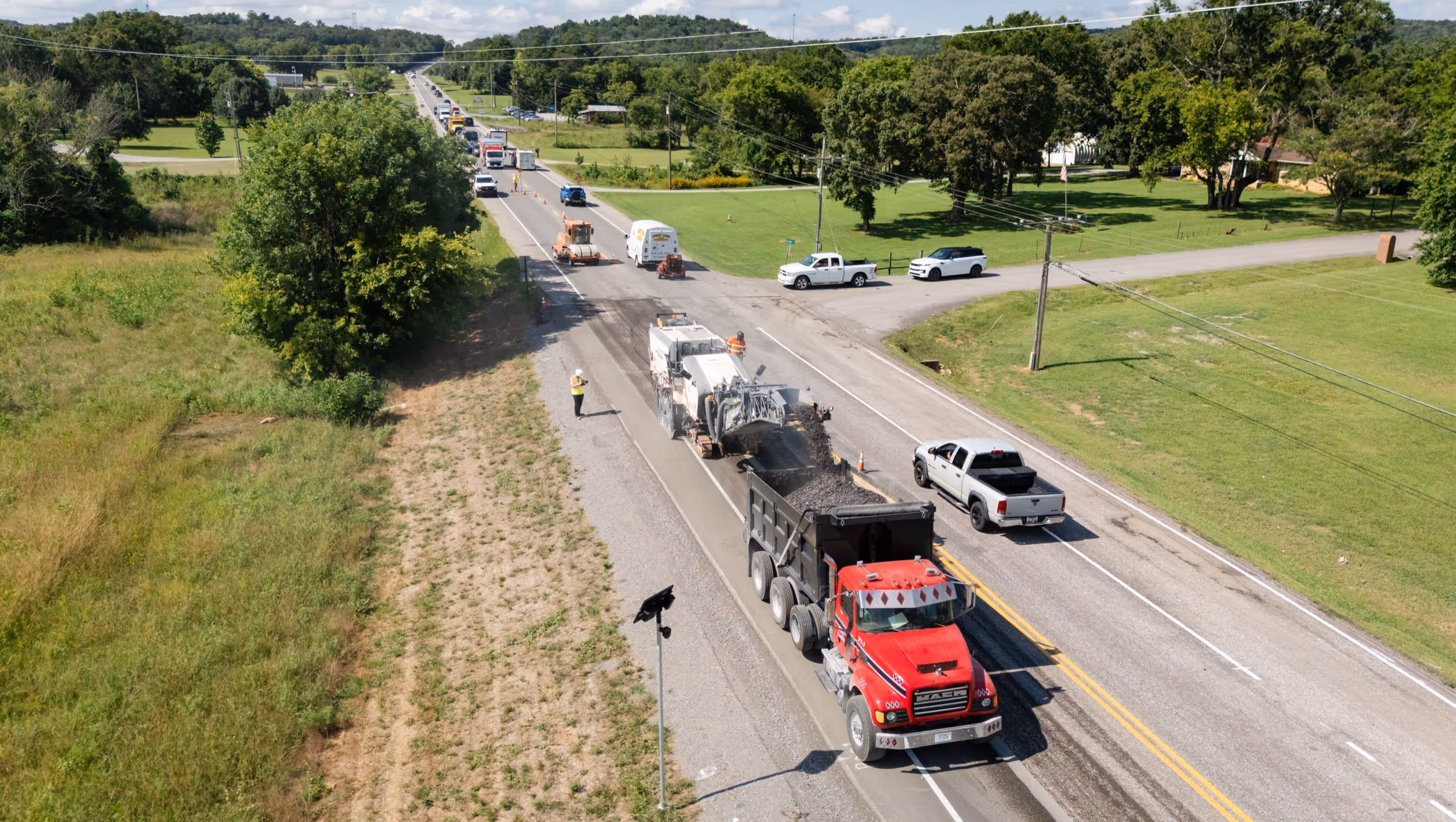 Road construction scene with a red dump truck receiving asphalt from a paving machine on a two-lane rural road.
