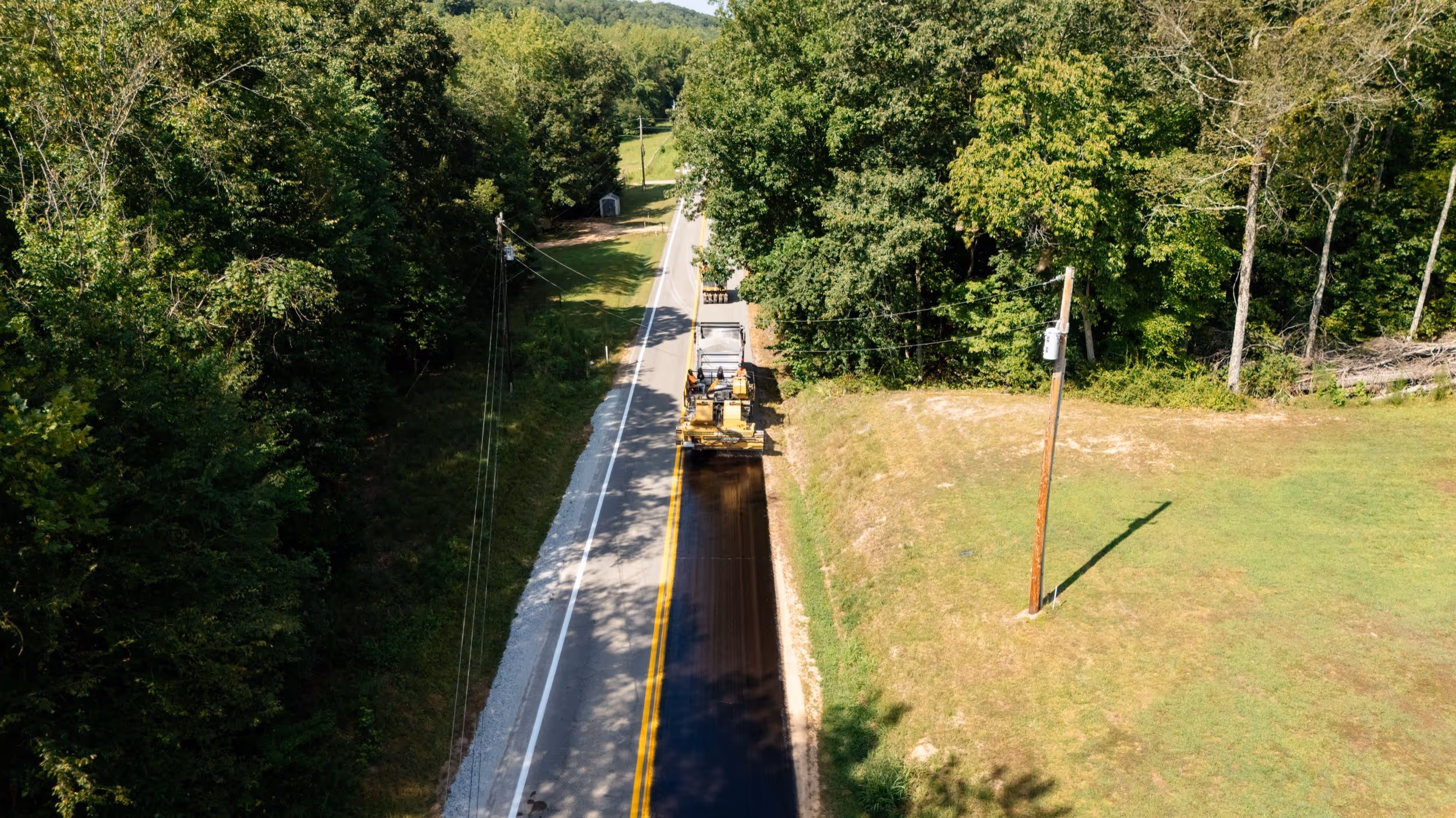 A road construction vehicle paving a narrow two-lane road surrounded by trees and grassy areas.