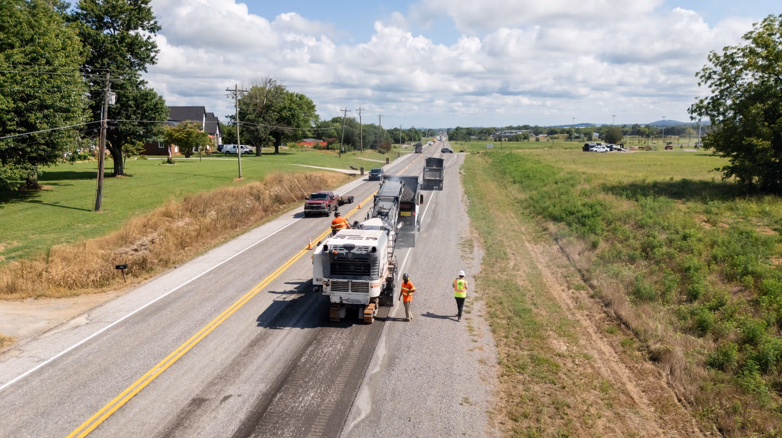 Road construction crew milling pavement on a rural highway with trucks collecting asphalt.