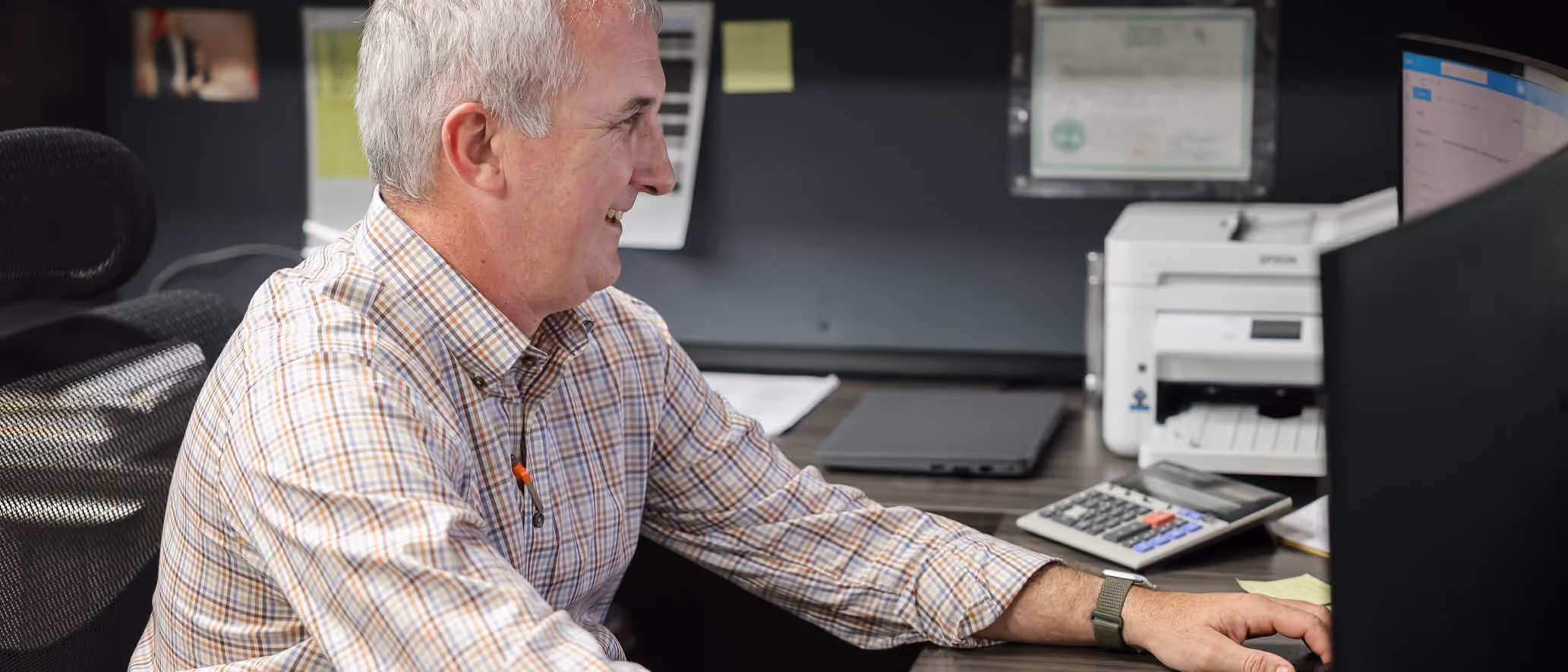 Middle-aged man in a plaid shirt smiling while working at a desk with a computer, calculator, printer, and laptop.