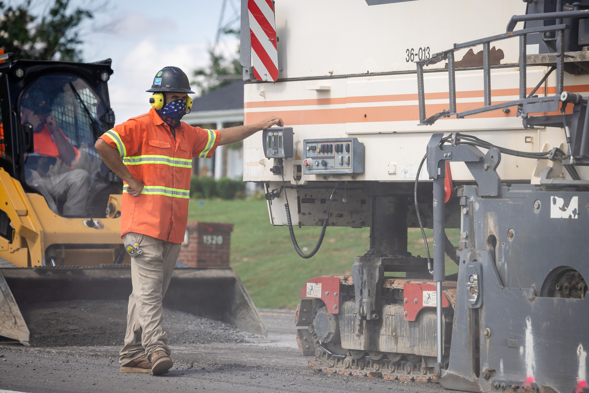 Construction worker in orange safety shirt and helmet with earmuffs standing next to heavy machinery on a roadwork site.