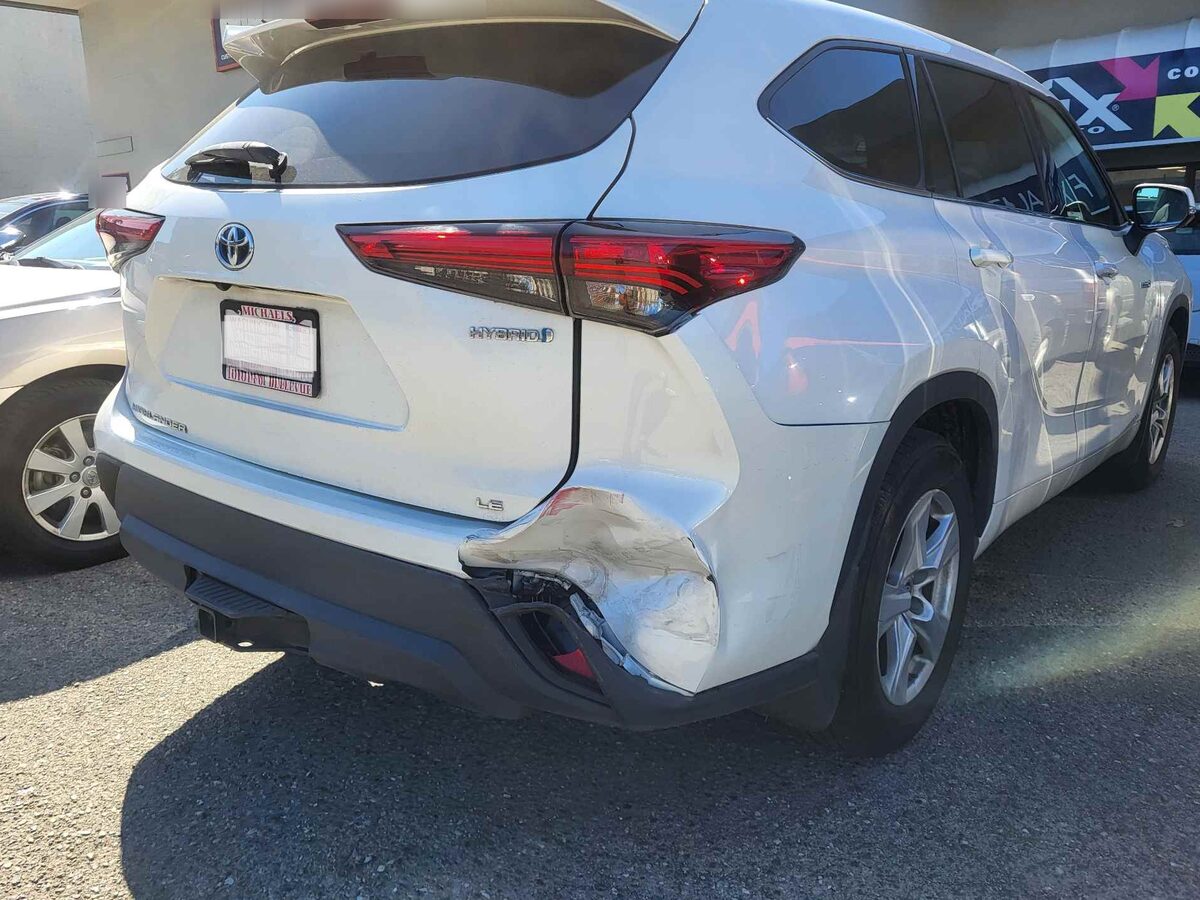Rear of a white Toyota Highlander Hybrid as it arrived, the bumper cover torn open and a crease running up into the quarter panel