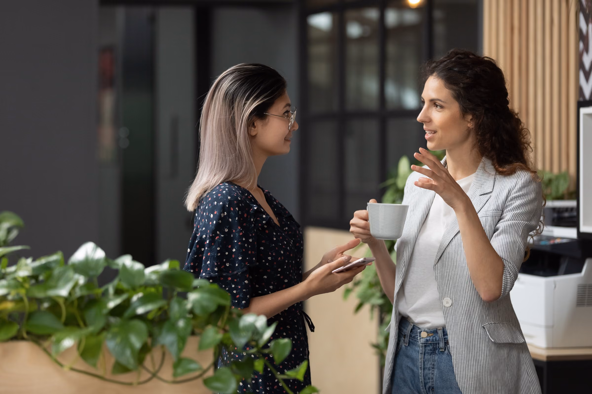 Two women talking in an office, one holding a coffee cup, with leafy plants in the foreground