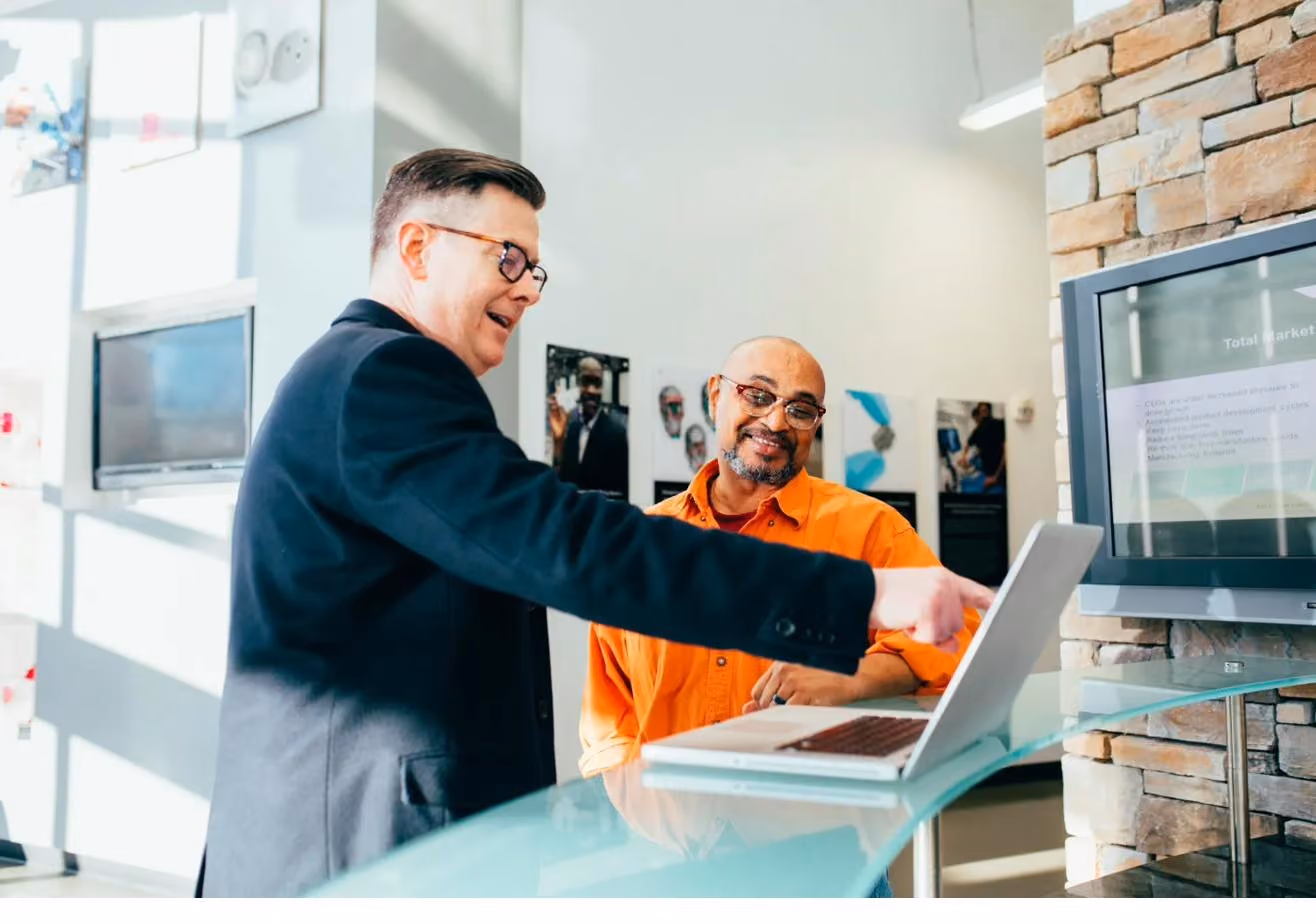 A man in a suit and glasses leaning over a laptop at a counter, smiling alongside a colleague in a hi-vis orange shirt