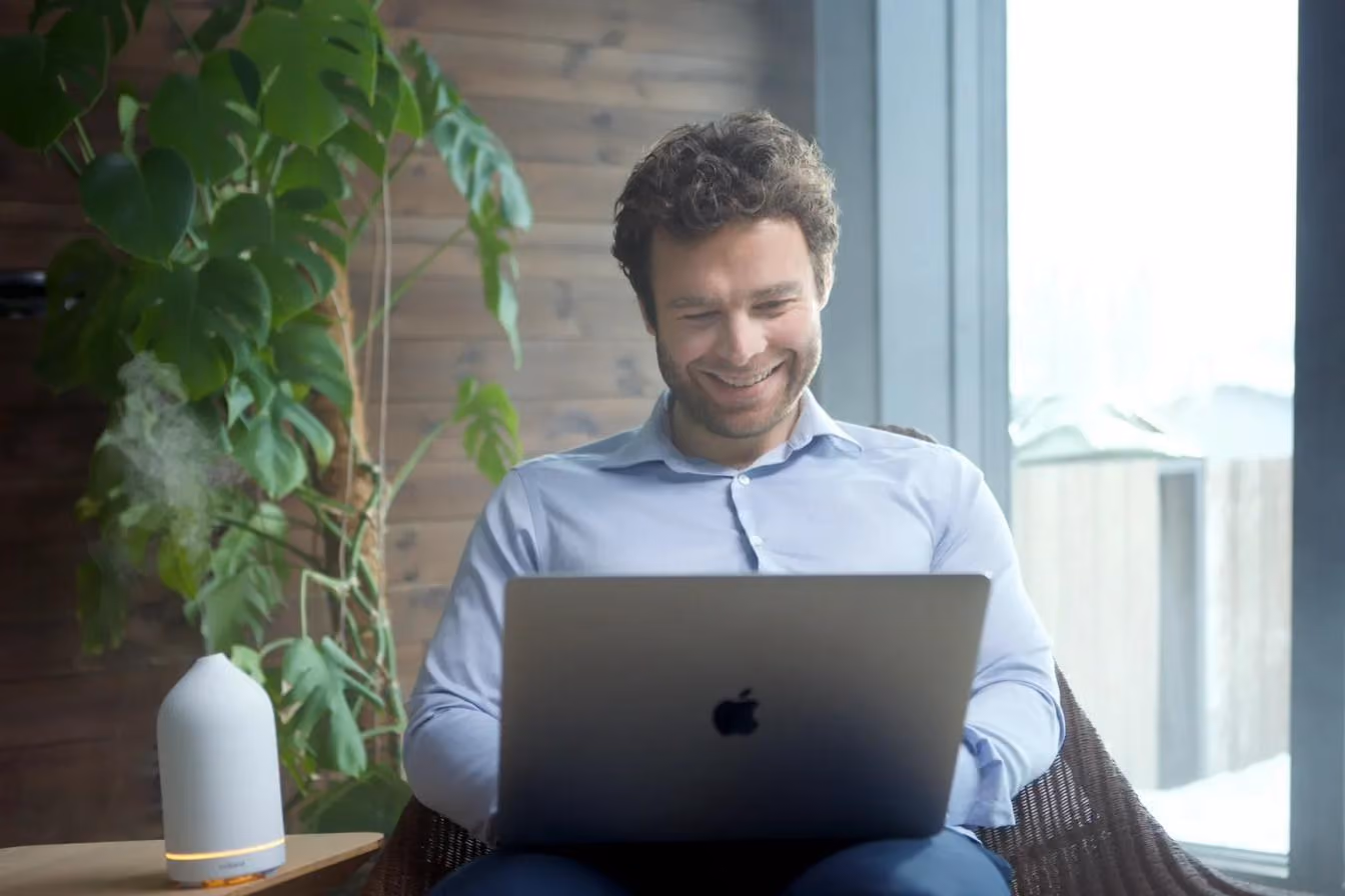 A man in a blue shirt smiling as he works on a laptop in an armchair beside a window and a large plant