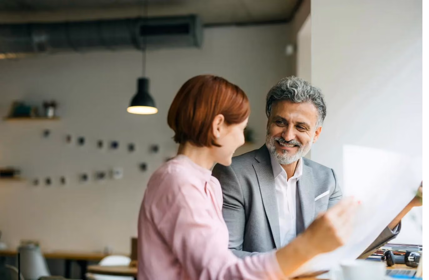 A woman and a grey-haired man in a blazer smiling at each other over a document in a bright, casual meeting space