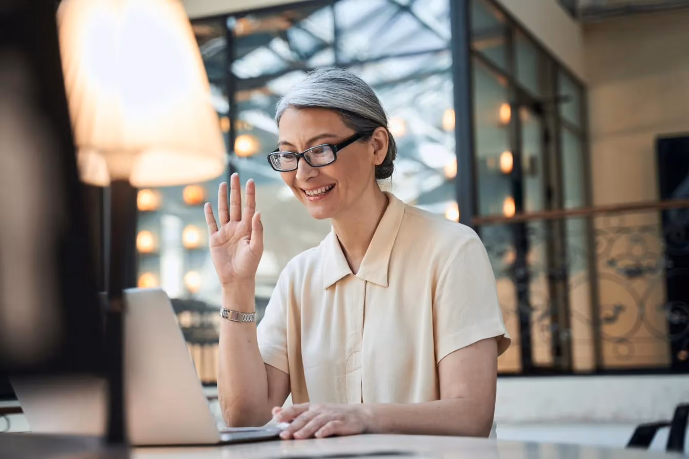A woman with grey hair and glasses smiling and waving at her laptop screen during a video call