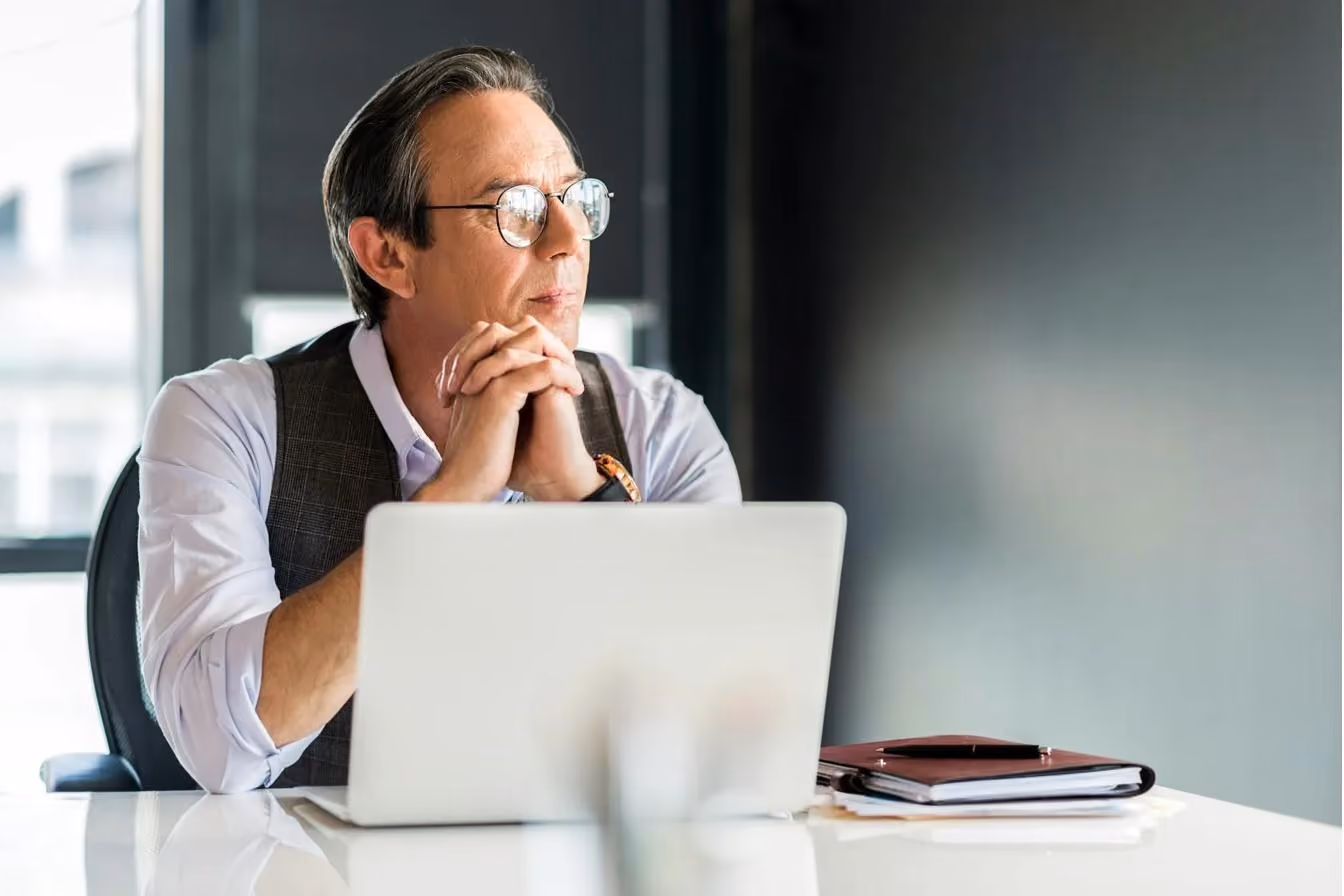 One lone executive with glasses, brown hair and a vest looking out a window lost in thought.