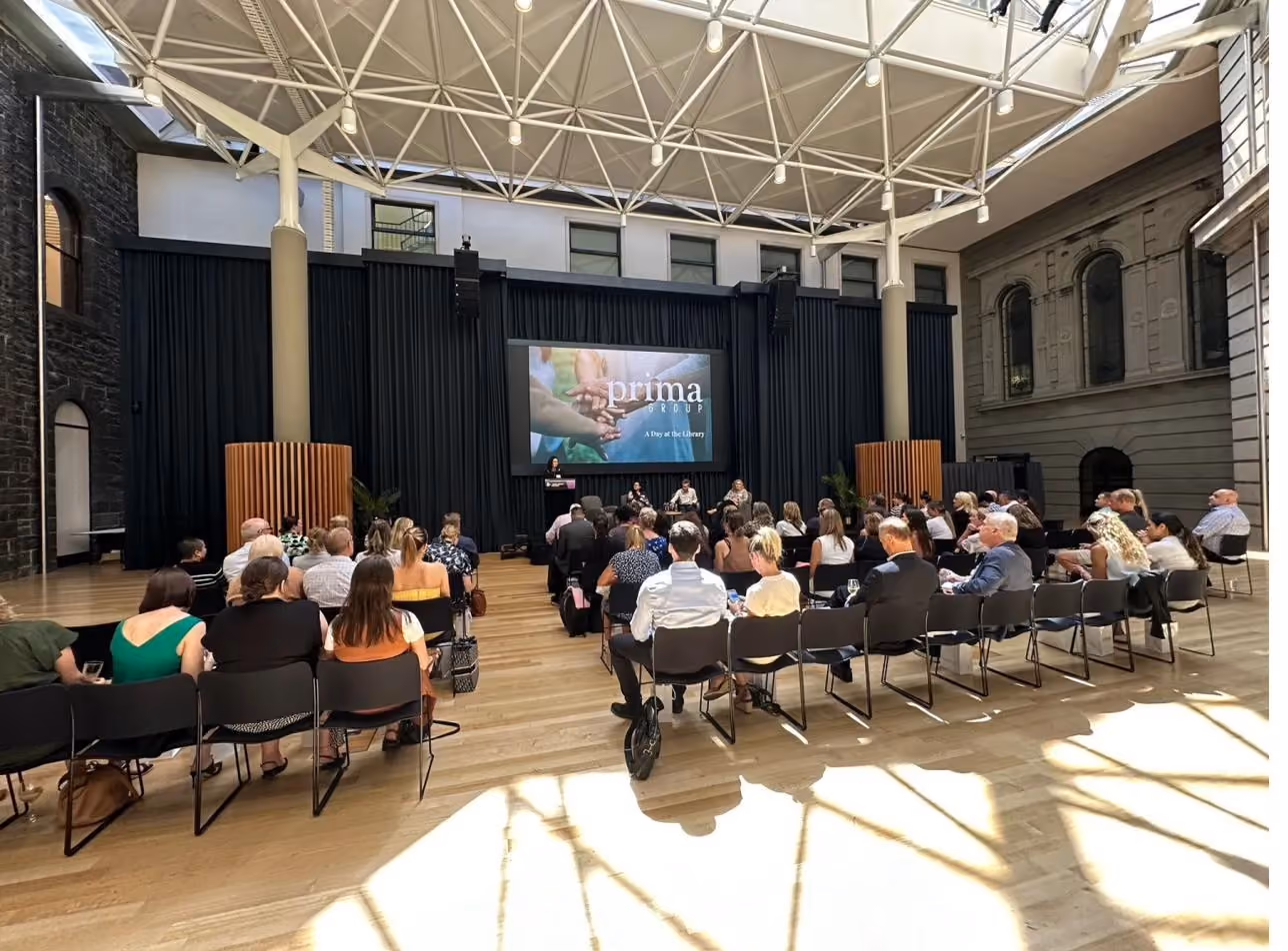 Many people seated in a large, well lit, white amphitheatre for Prima’s “day at the library” presentation.