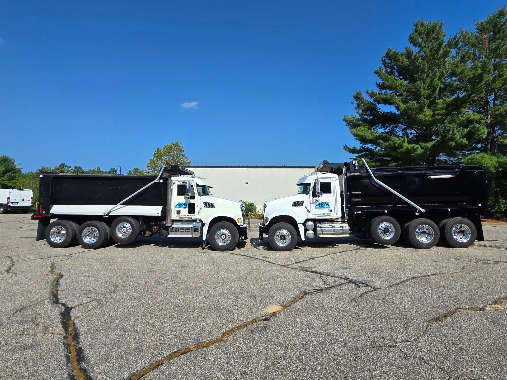 Two white dump trucks with black beds facing each other on a cracked asphalt surface with trees and a building in the background under a clear blue sky.