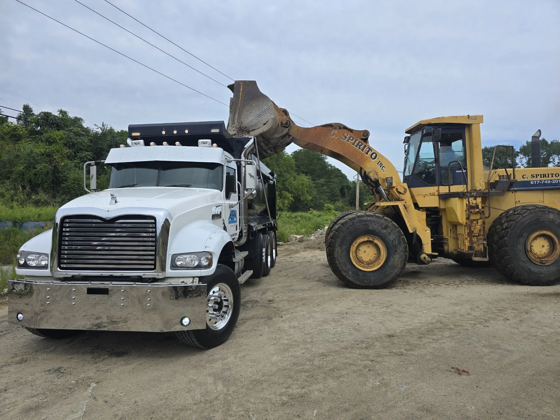 White dump truck parked on a dirt surface with a yellow front loader beside it, the loader's bucket raised over the truck bed.