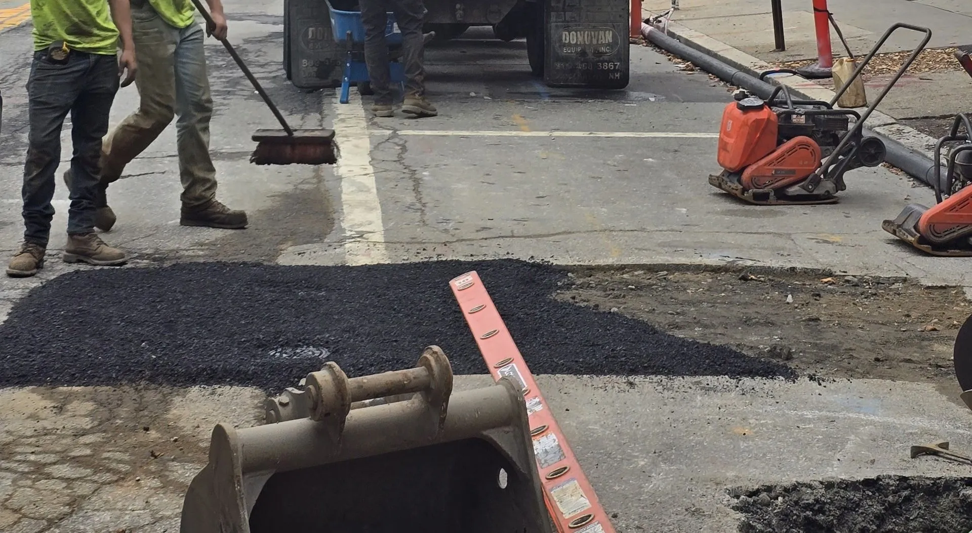 Two workers in boots and work pants repairing a road with fresh asphalt, one sweeping the surface while various paving equipment and a truck are nearby.