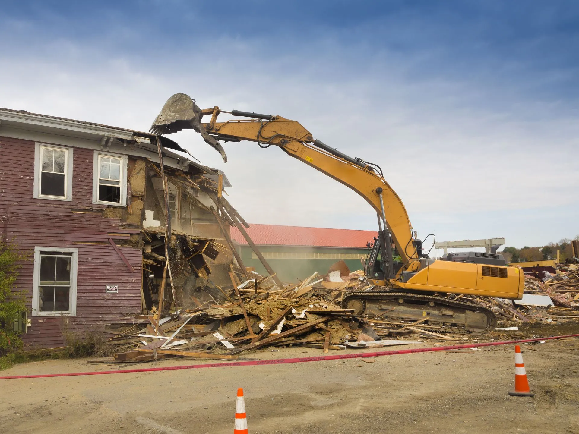 Yellow excavator demolishing a two-story wooden building with red siding, scattering debris around, with orange traffic cones and caution tape in the foreground.