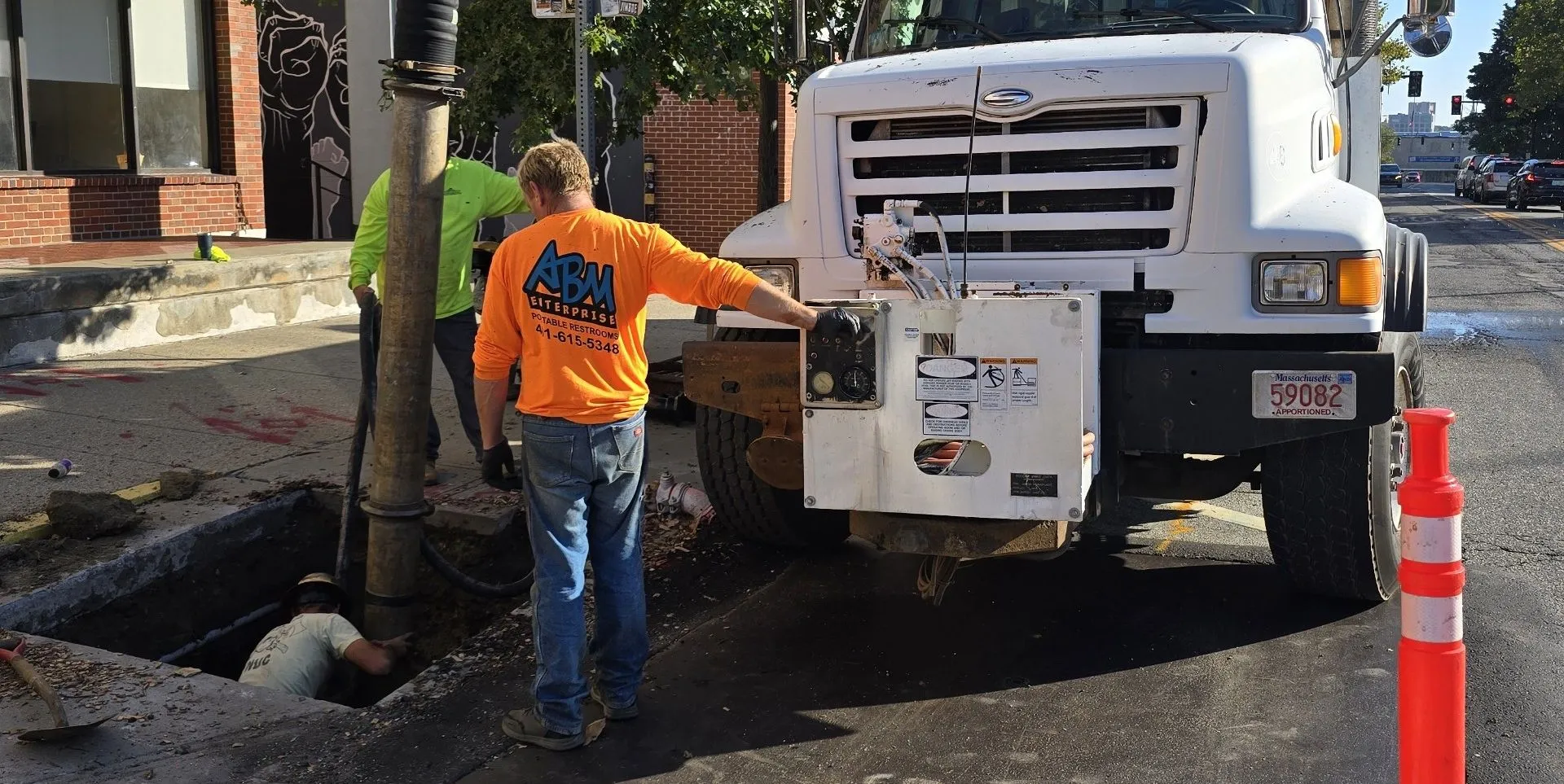 Three workers performing street maintenance by a manhole, one standing inside the hole, another in an orange ABM Enterprise shirt holding a control panel on a white truck, and another in a neon yellow shirt assisting.