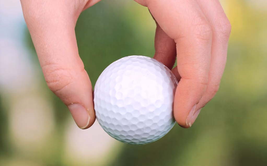 Close-up of a hand holding a white golf ball with a blurred green background.
