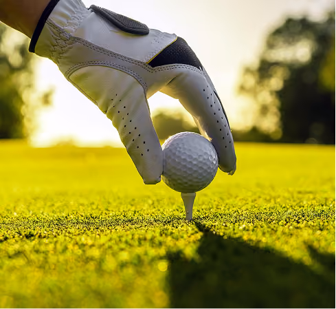 Hand wearing a white golf glove placing a golf ball on a tee on green grass with a blurred background in sunlight.