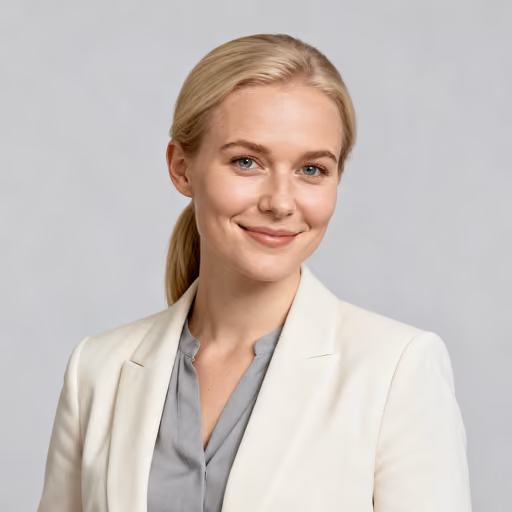 Smiling young woman with blonde hair tied back, wearing a white blazer and light gray blouse, against a plain gray background.