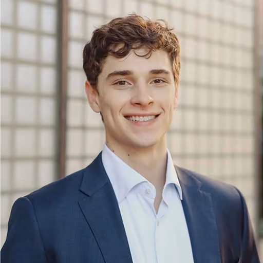 Young man with short curly brown hair wearing a white shirt and dark blue blazer, smiling against a blurred background of light-colored tiles.