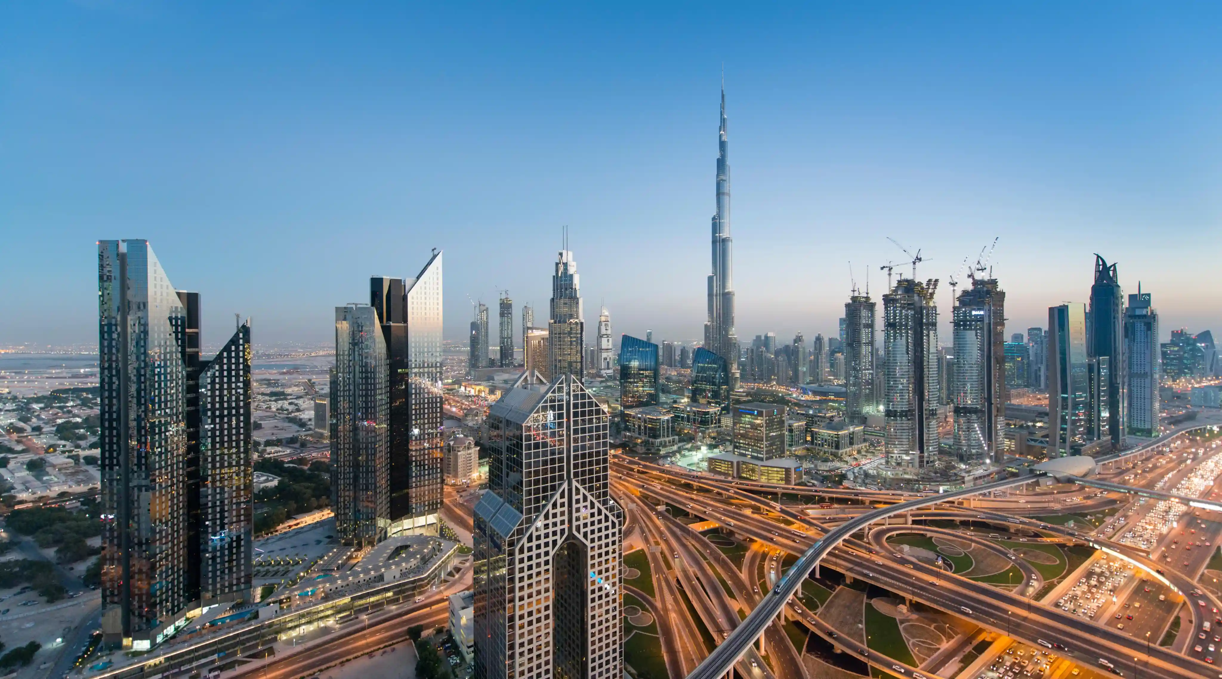 Skyline of a modern city at dusk with numerous high-rise buildings including the tallest skyscraper, intricate highway interchanges with flowing traffic, and construction cranes visible in the background under a clear sky.