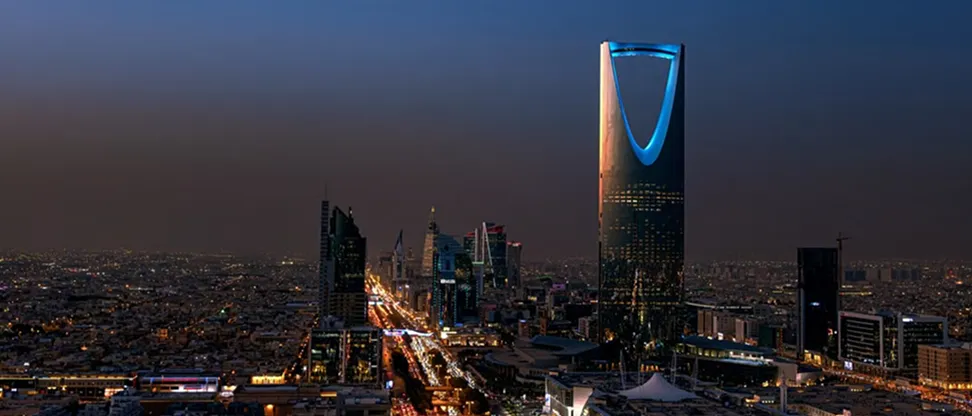 City skyline at dusk featuring the illuminated Kingdom Centre tower with its distinctive curved top, surrounded by other skyscrapers and streets filled with lights.