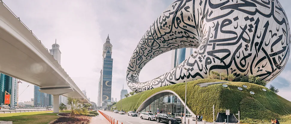 Modern urban scene featuring the Museum of the Future in Dubai with its elliptical structure covered in Arabic calligraphy, adjacent to a green landscaped base and a nearby elevated metro track with city skyscrapers in the background under a partly cloudy sky.