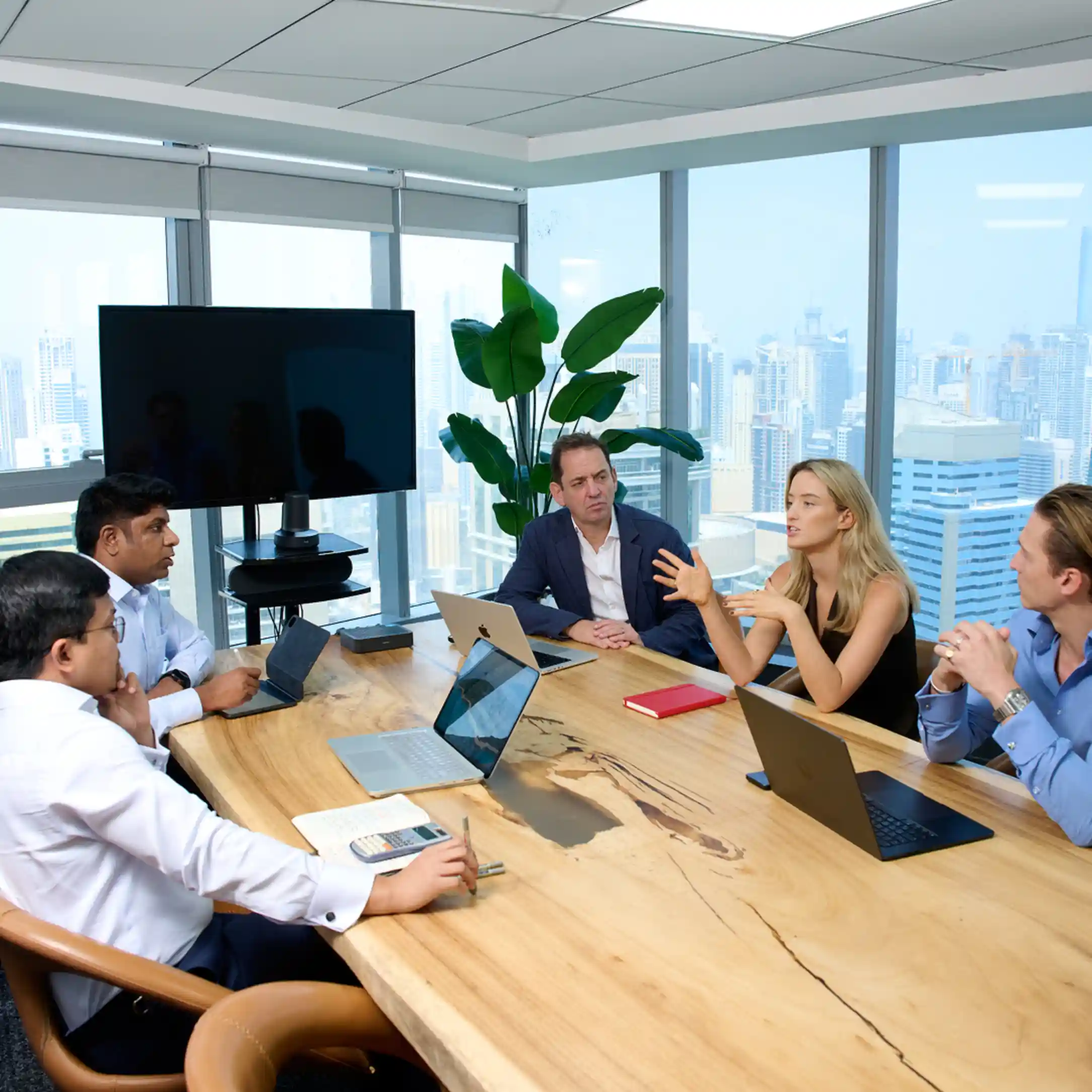 Five business professionals seated around a large wooden conference table in a modern office with floor-to-ceiling windows showing a cityscape, engaged in discussion with laptops and notebooks in front of them, and a large screen and indoor plant visible in the background.