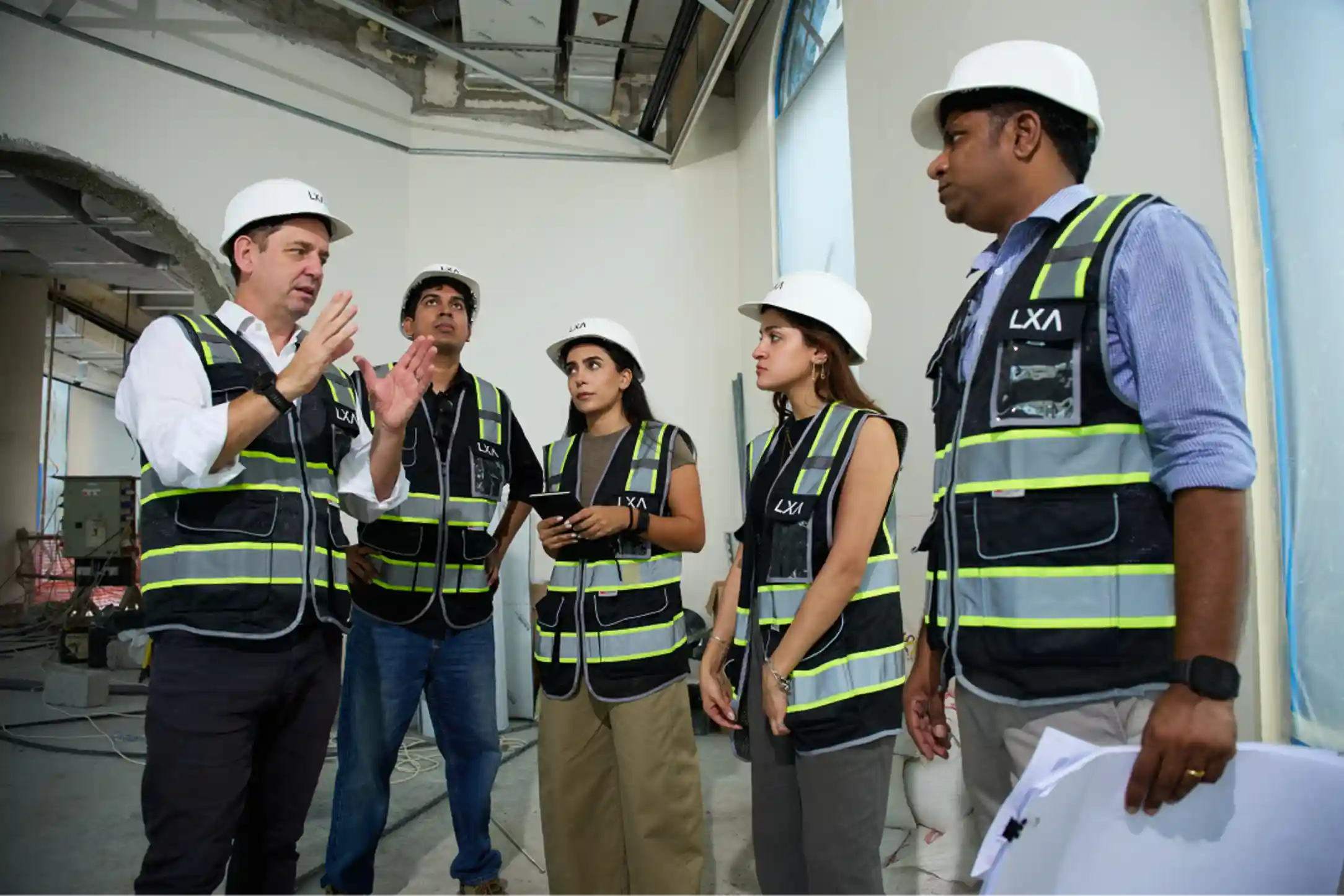 Five construction professionals wearing white safety helmets and black reflective vests stand inside an unfinished building, with one man gesturing as he speaks to the group while others listen attentively, holding a tablet and documents.